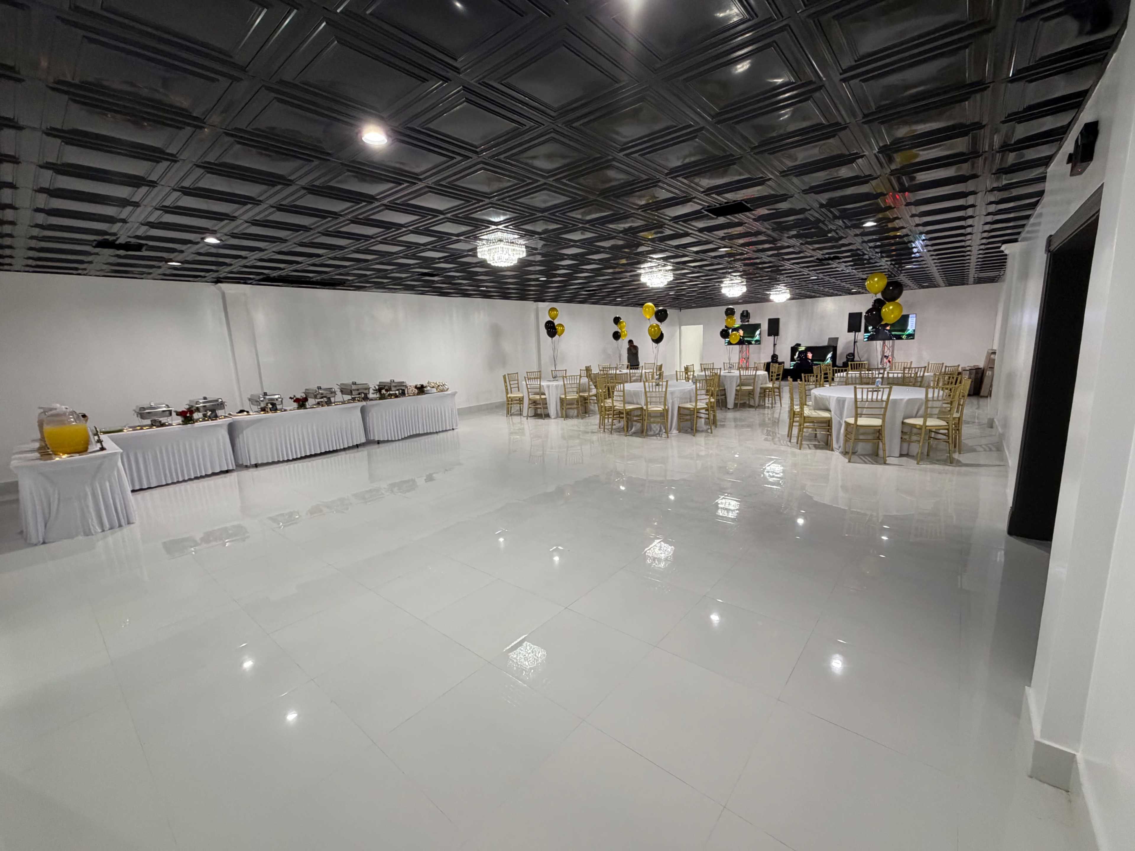 The image shows a spacious event room with white walls, a black ceiling, tables with gold chairs set for dining, and a buffet table along one side.