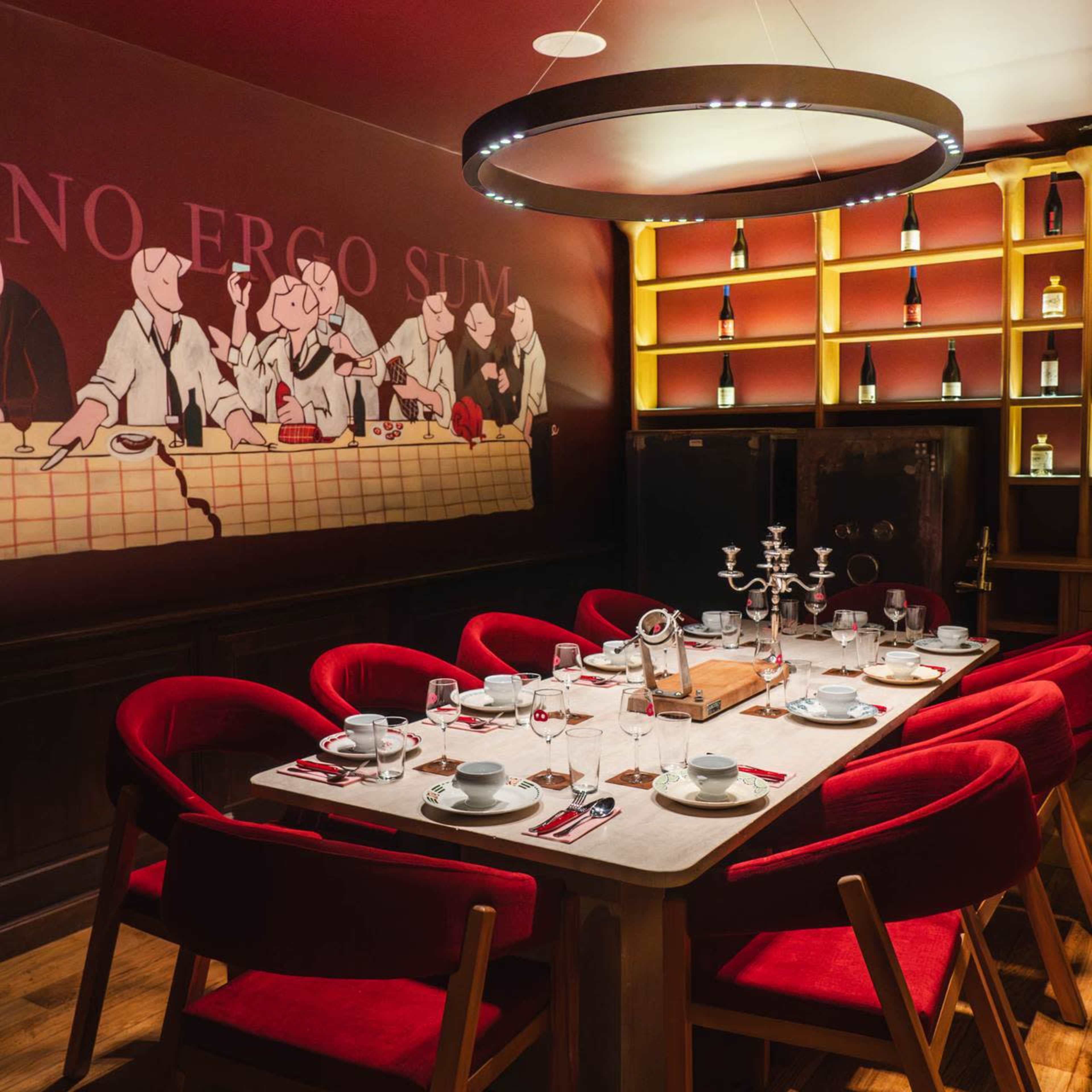 The image shows a dining table set with plates and glasses in a restaurant featuring a mural of people dining on the wall and illuminated shelves displaying bottles.