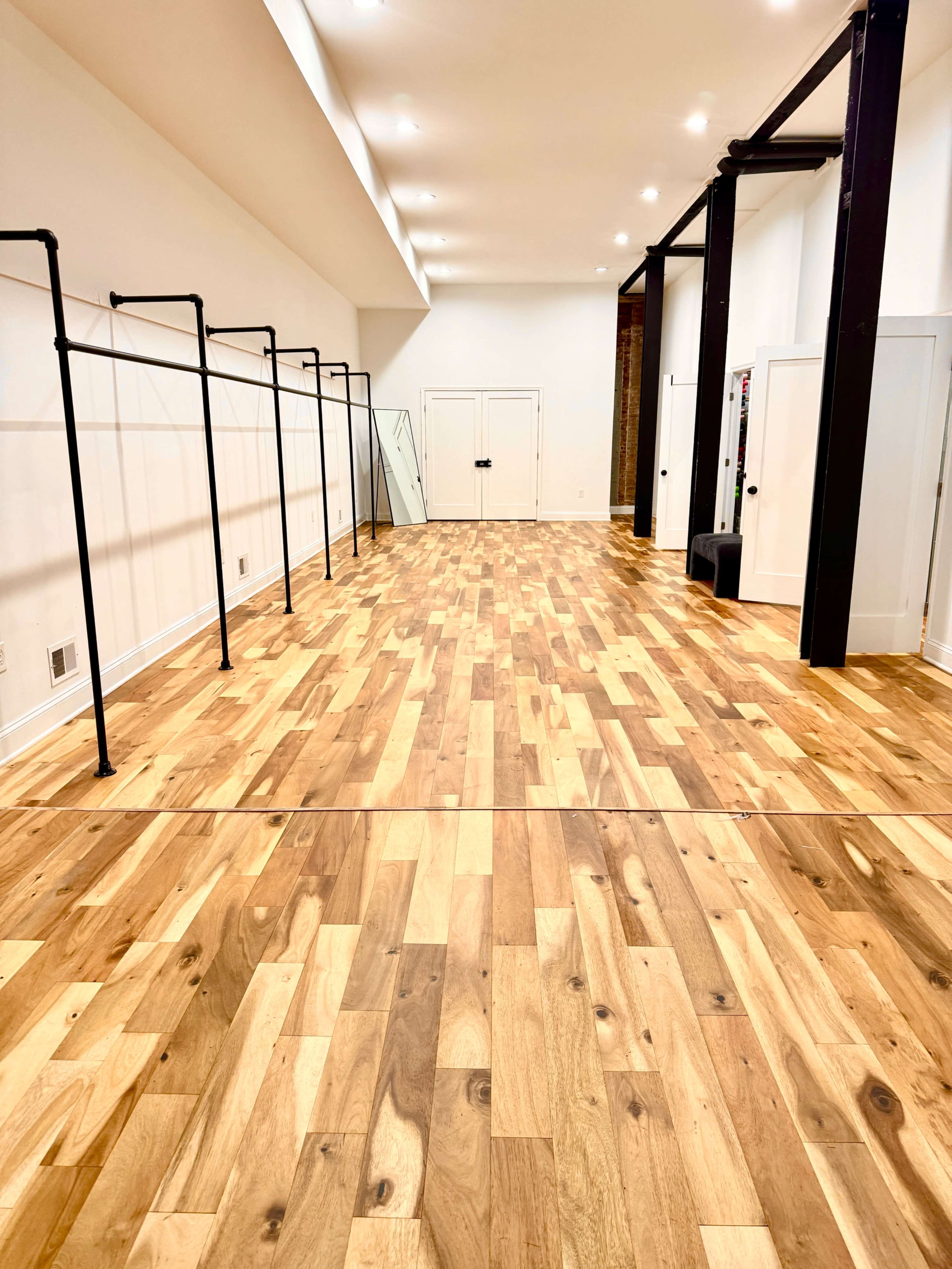 The image shows a spacious, well-lit room with light wooden flooring, equipped with black clothing racks along one wall and a mirror in the corner.