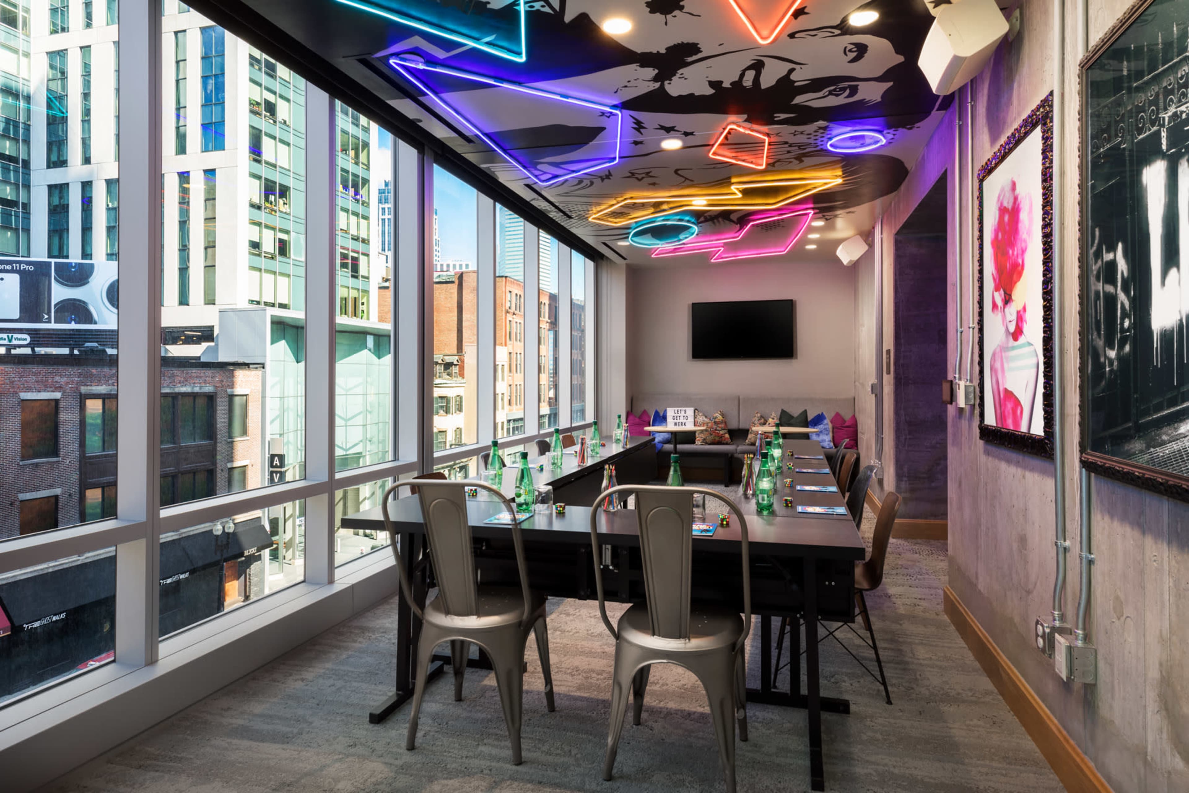 A modern conference room features large windows with city views, a long dark table, and colorful neon lighting on the ceiling.