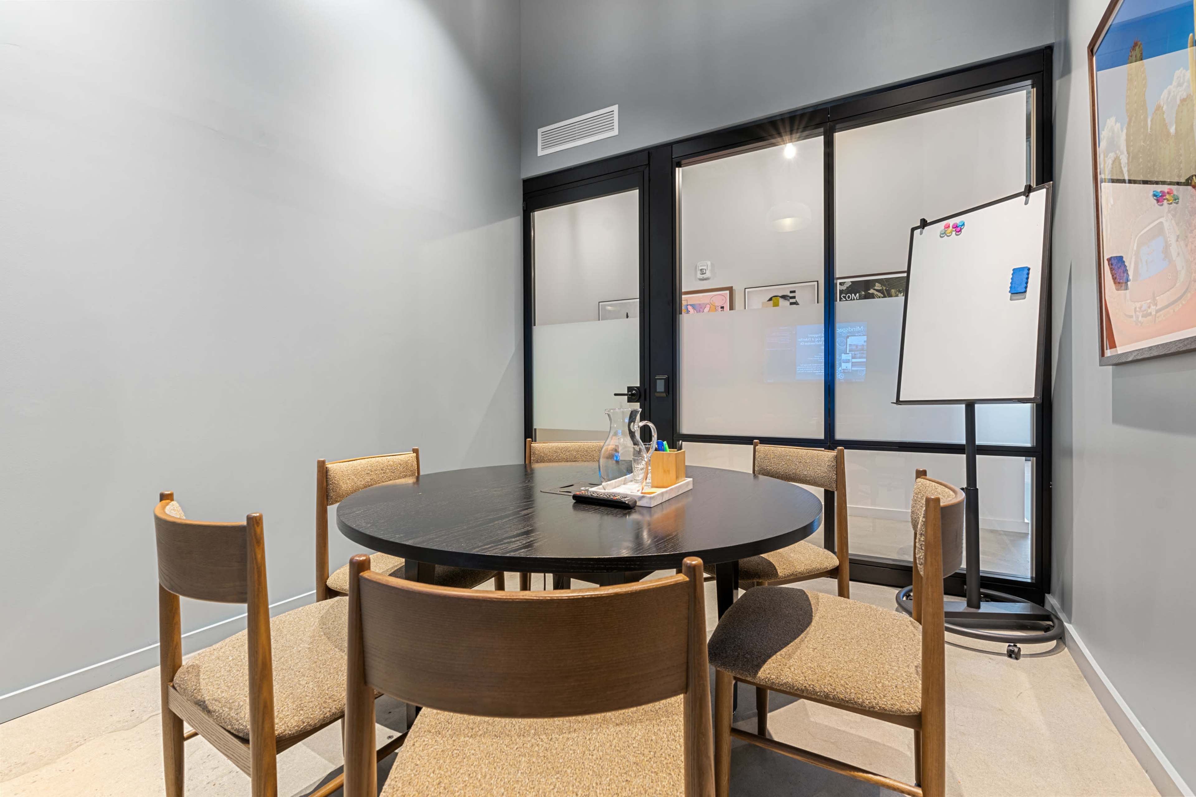 A conference room features a round table surrounded by four wooden chairs, with a glass partitioned space and a whiteboard in the background.