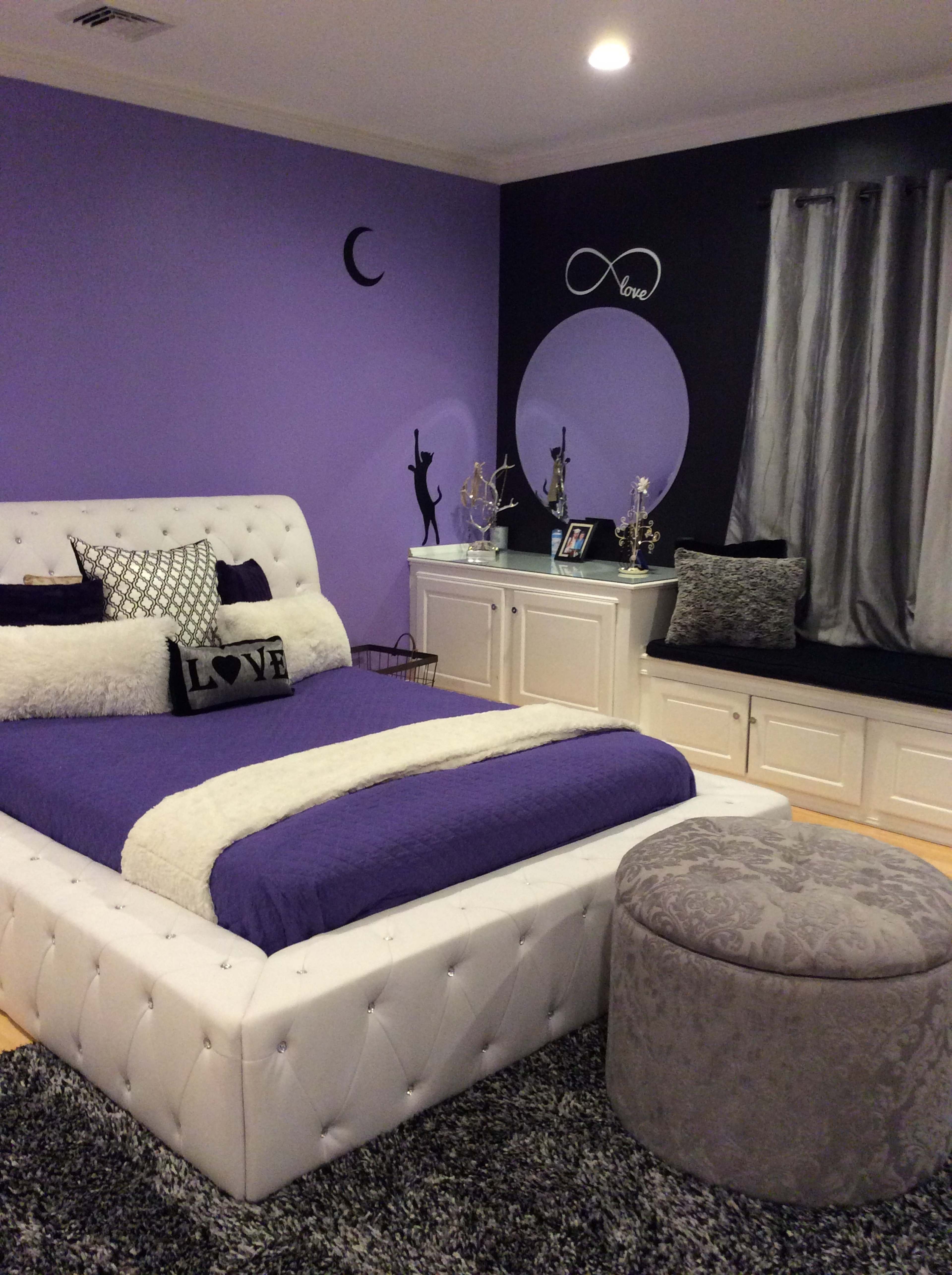 A modern bedroom with purple walls, a white tufted bed with purple bedding, and a decorative black and white accent wall featuring a circular mirror.