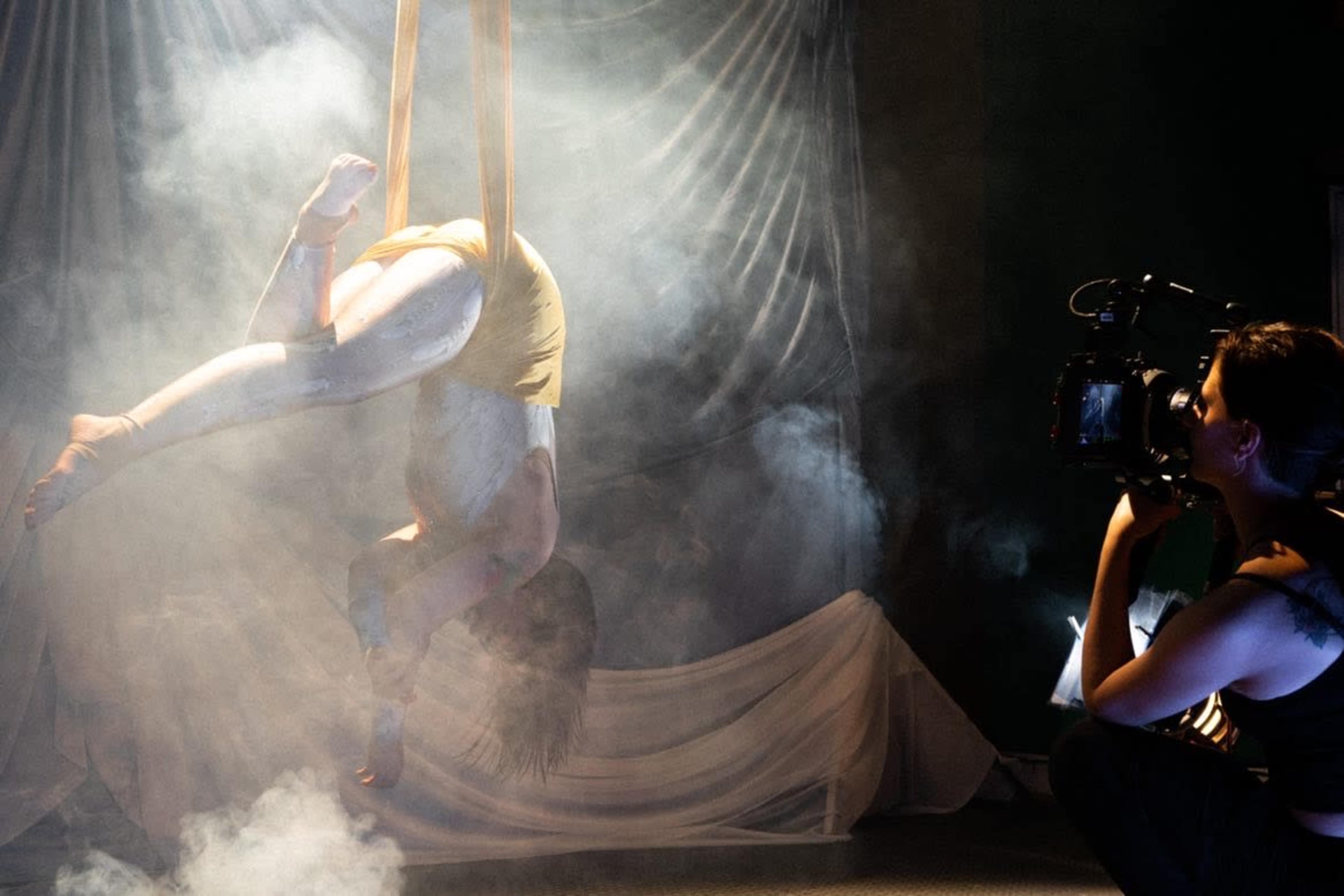 A performer is suspended in the air using fabric while a woman captures the scene with a camera, all amidst a backdrop of smoke and draped fabric.