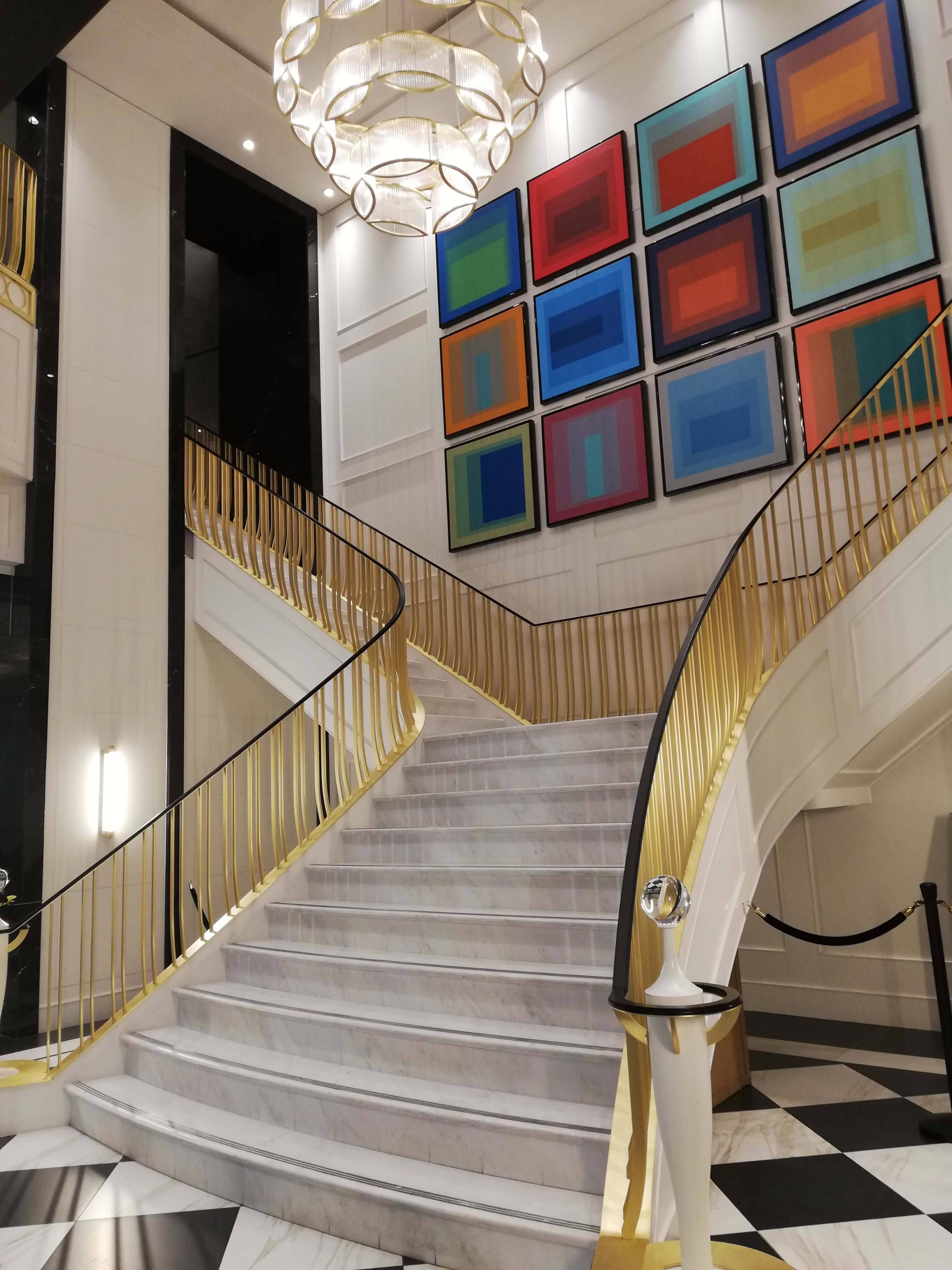 The image shows a grand staircase with a gold railing, leading upward in a well-lit space adorned with colorful framed artwork on the wall.