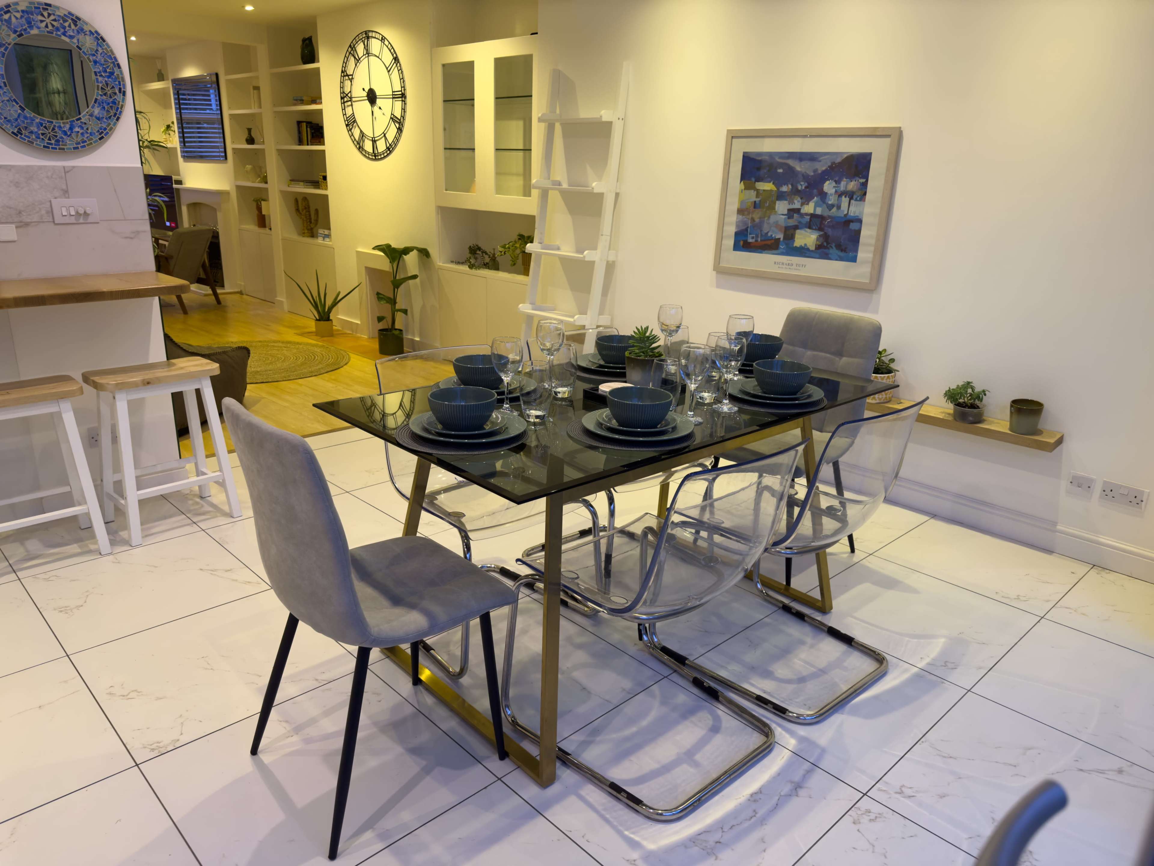 A modern dining room features a glass table set for eight, surrounded by gray chairs, with a clock and artwork displayed on the walls.