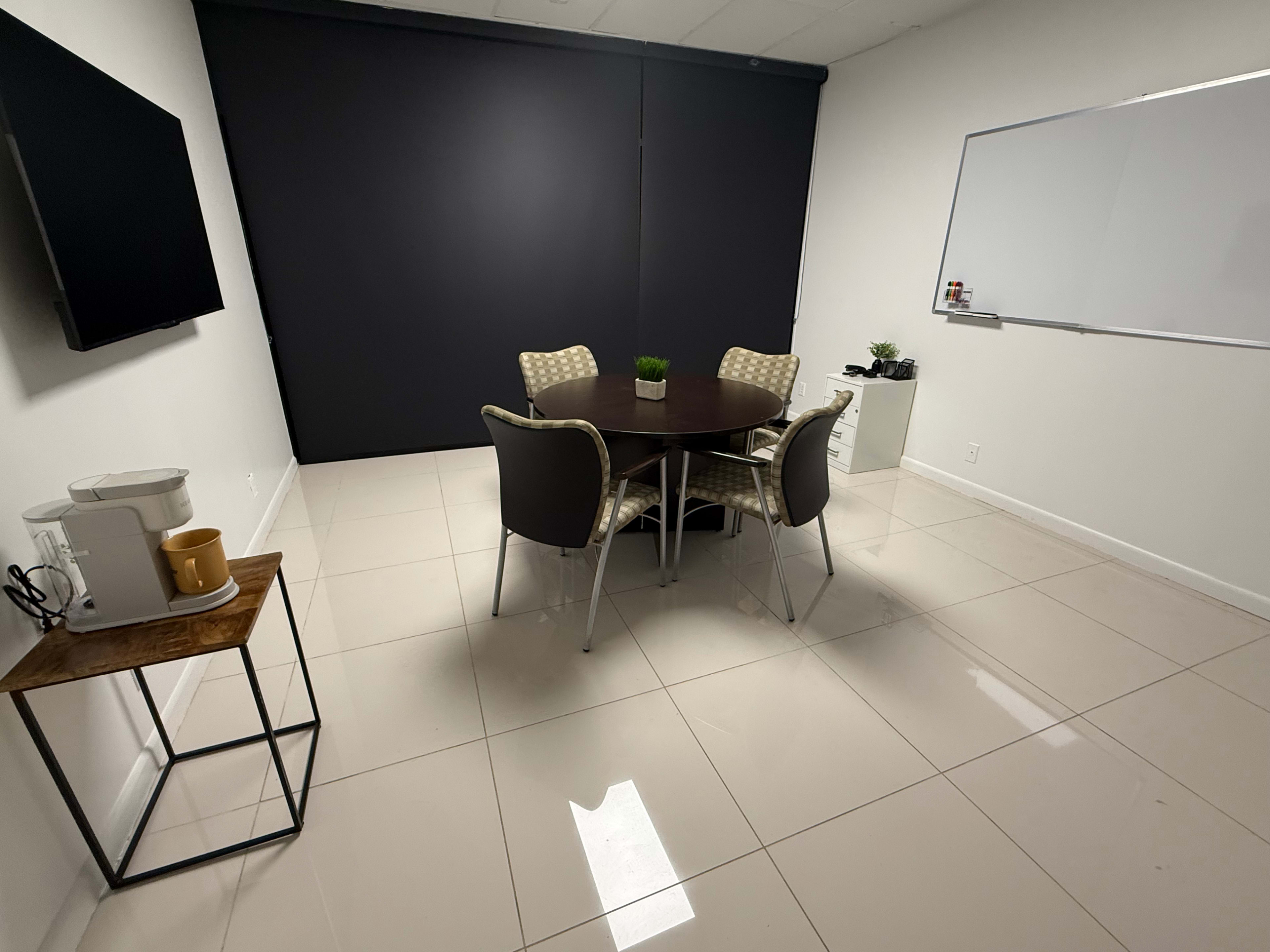 A meeting room features a round table surrounded by four chairs, a small side table with a coffee maker, a television mounted on the wall, and a whiteboard.