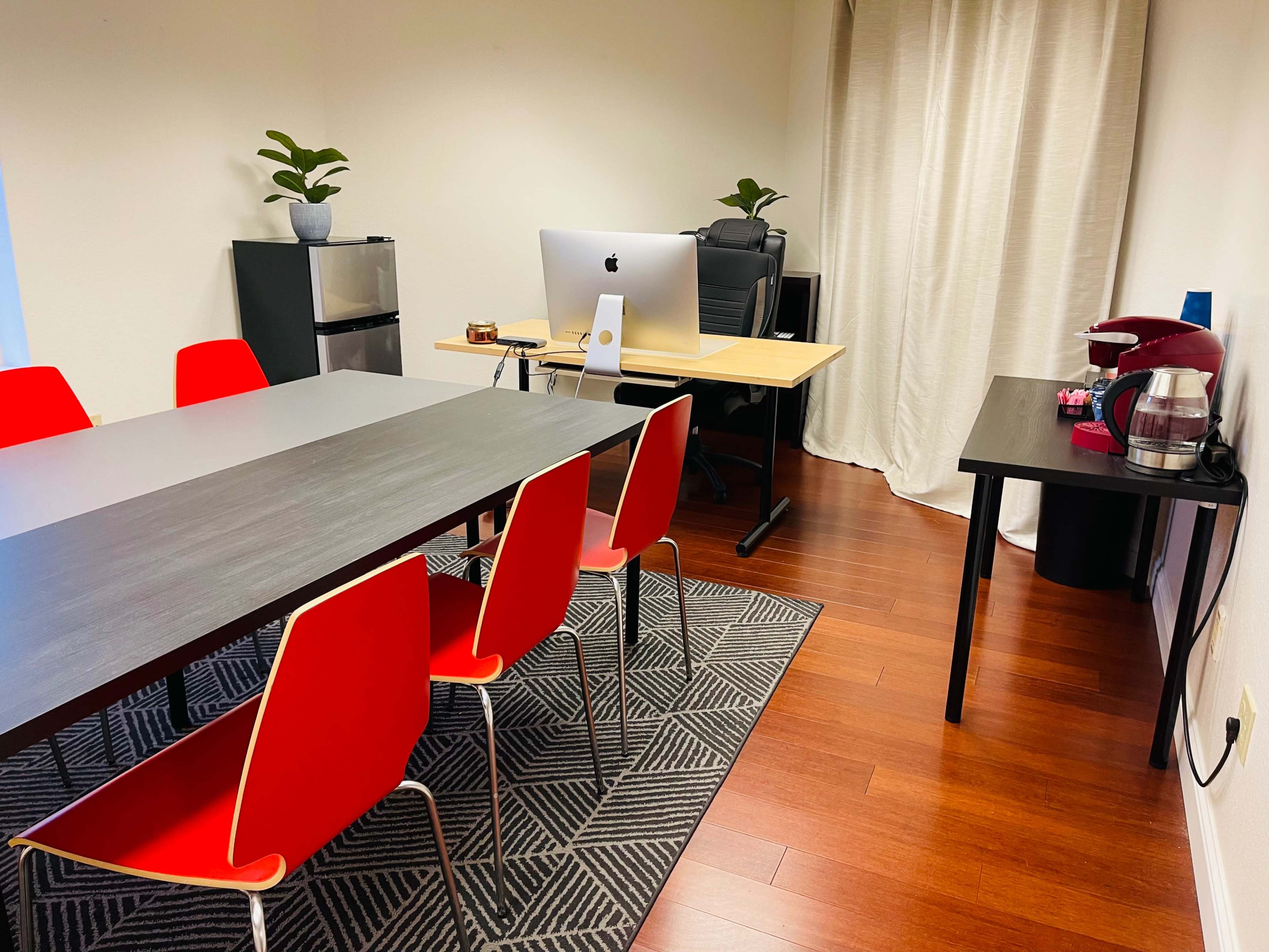 The image shows a modern office space with a conference table surrounded by red chairs, an Apple computer on a desk, and a coffee maker on a side table.