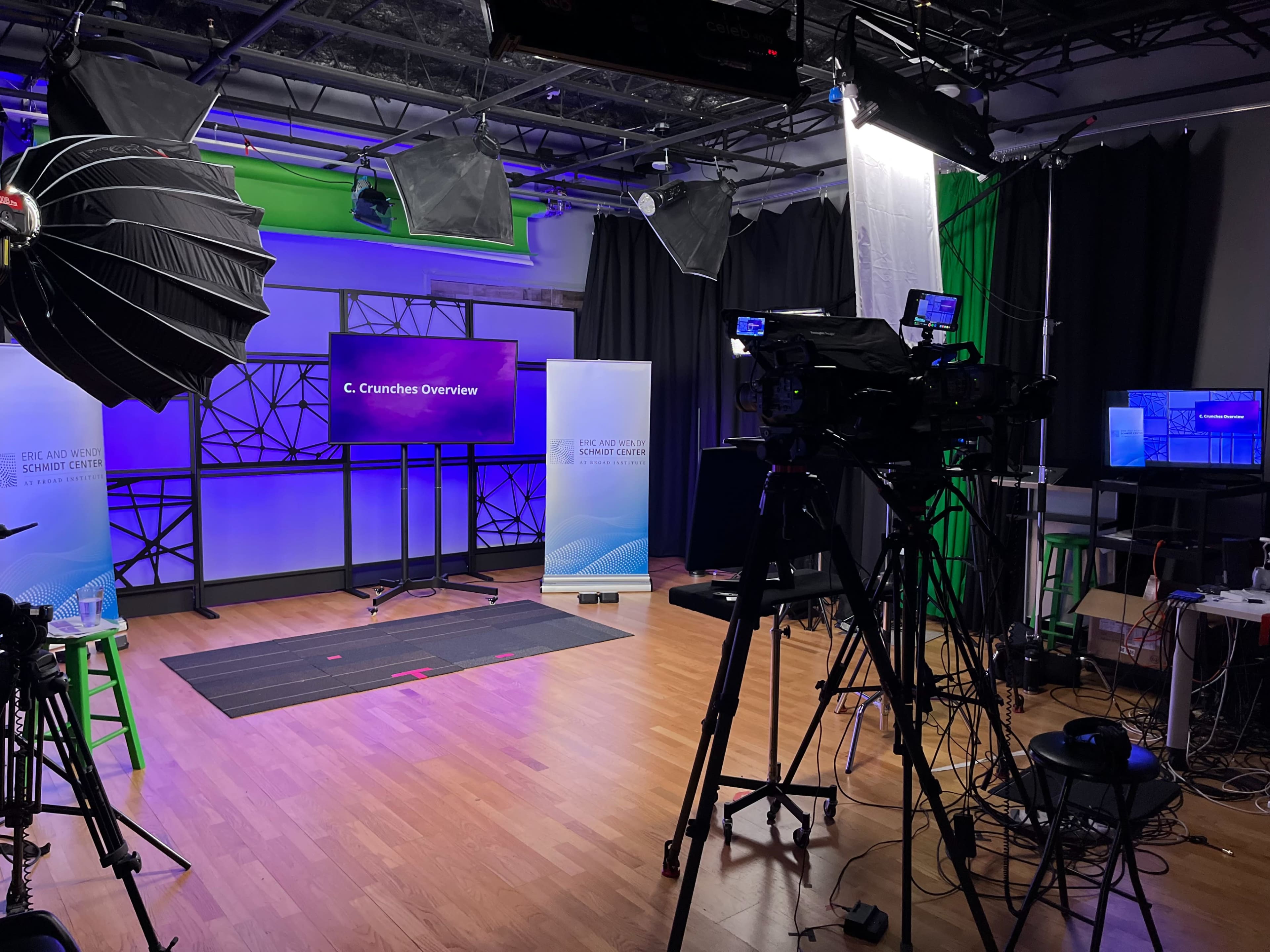 The image shows a television studio setup with multiple cameras, lights, and a large screen displaying "C. Crunches Overview" on a backdrop of green and black curtains.