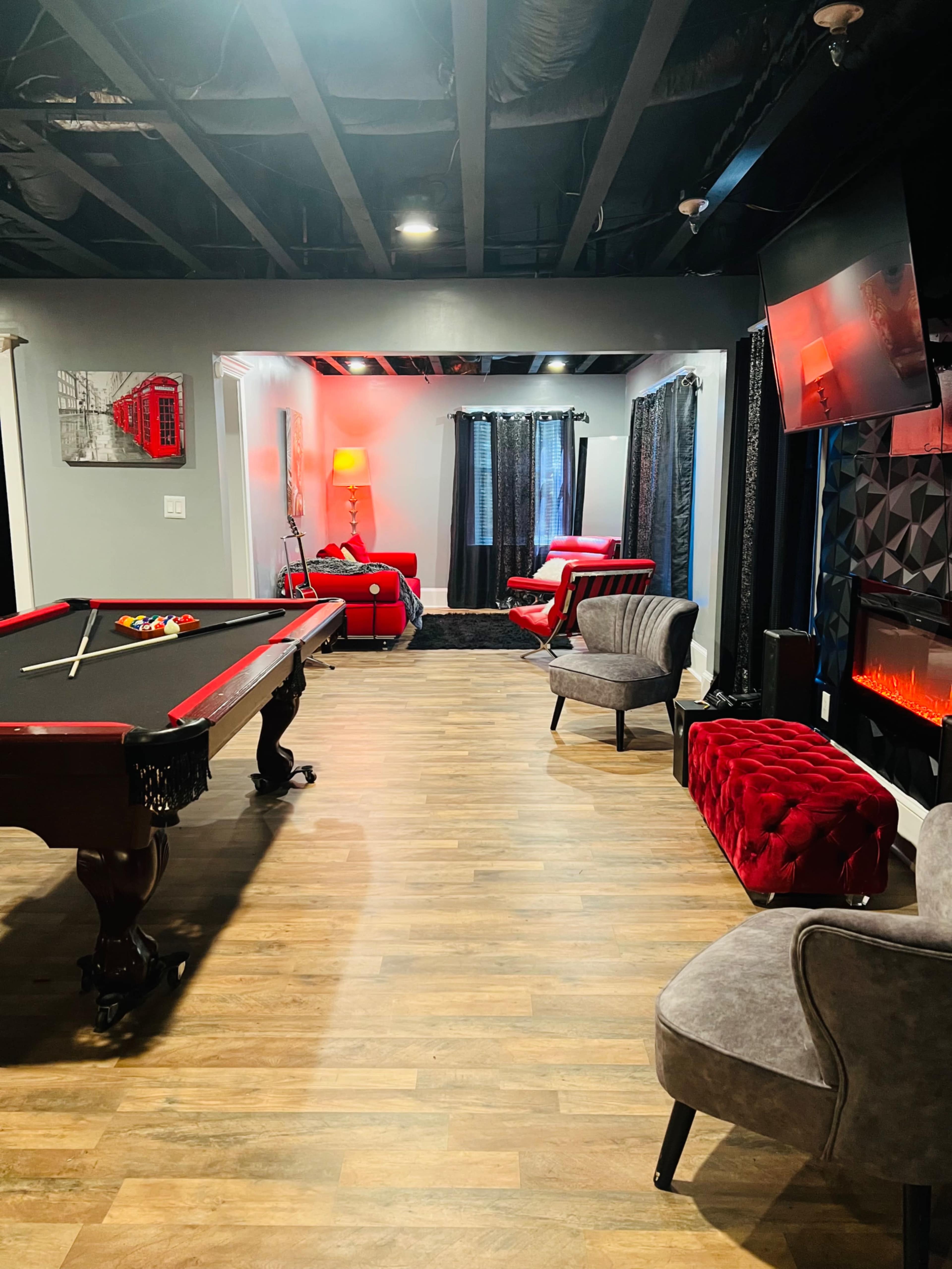 The image shows a recreation room featuring a pool table, a red velvet bench, a gray armchair, and a decorative electric fireplace under a television.