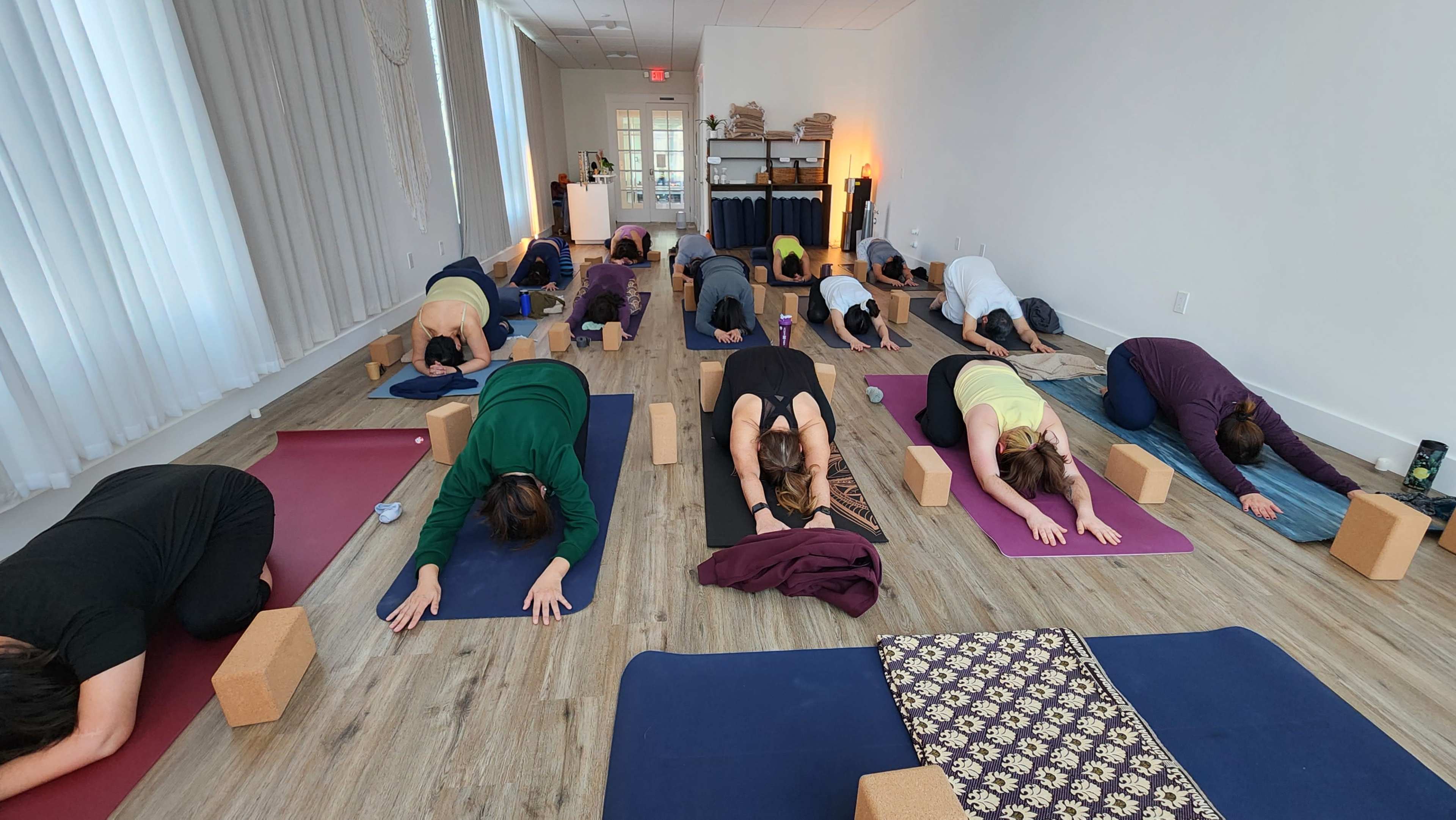 A group of people practices yoga in a bright studio, using blocks for support while in a resting pose.