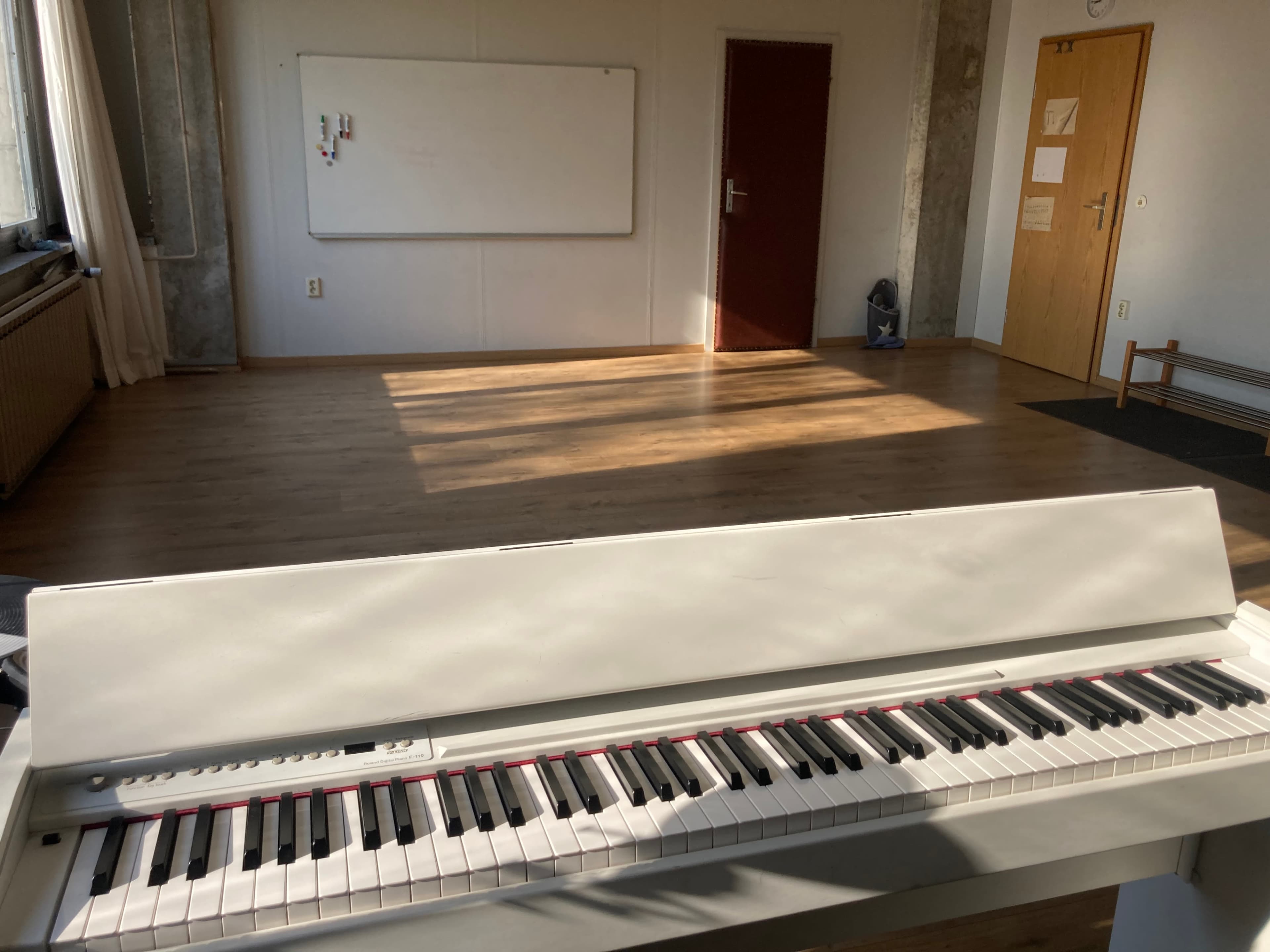 A white digital piano is positioned in the foreground of a sunlit room with wooden flooring and a whiteboard on the wall.