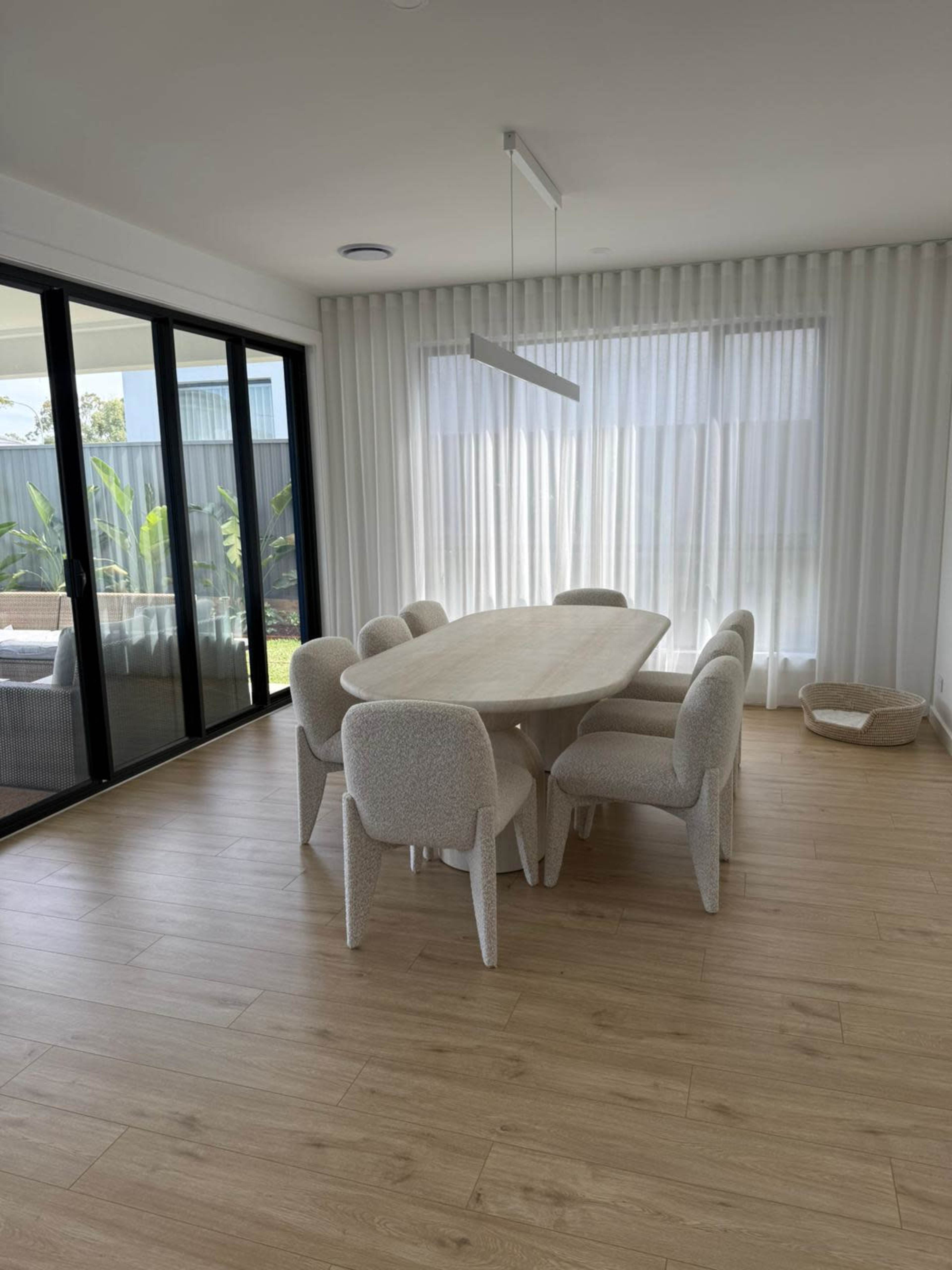 A modern dining area features a large oval table surrounded by upholstered chairs, with large windows letting in natural light.