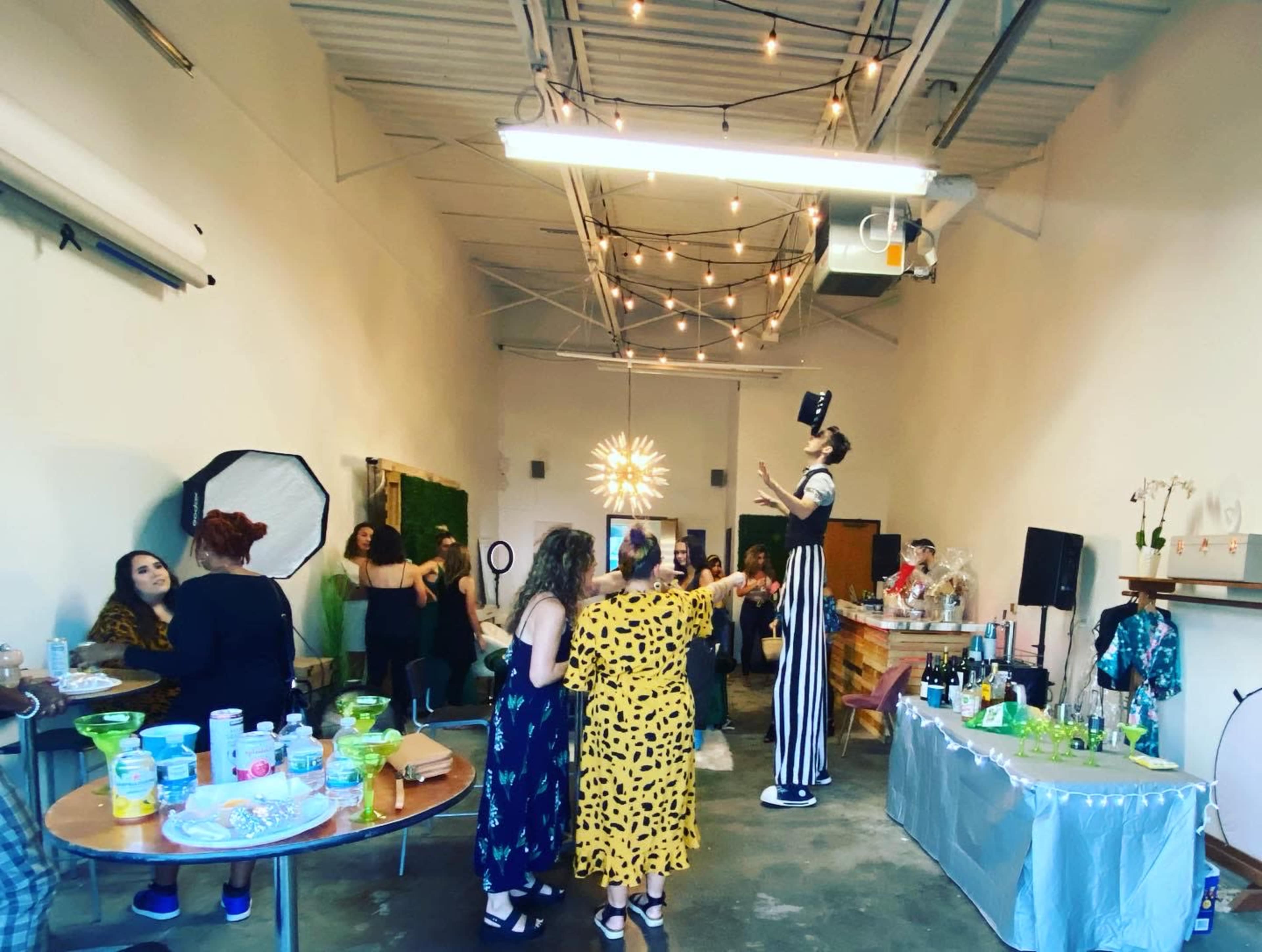 A lively gathering is taking place in a spacious venue with people mingling, tables of food and drinks, and a performer on stilts.