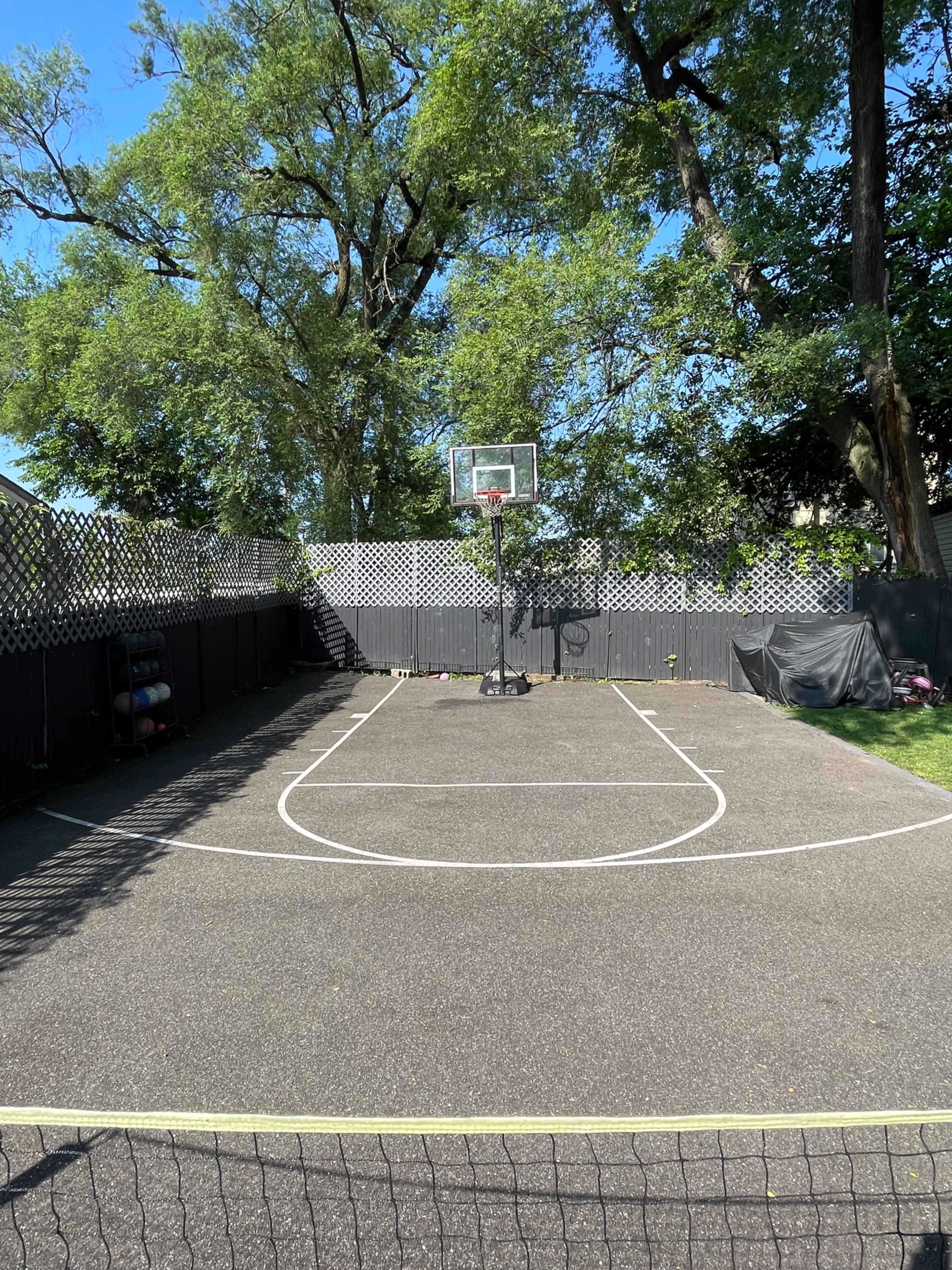 The image shows an outdoor basketball court surrounded by trees, featuring a hoop at one end and a fenced perimeter.