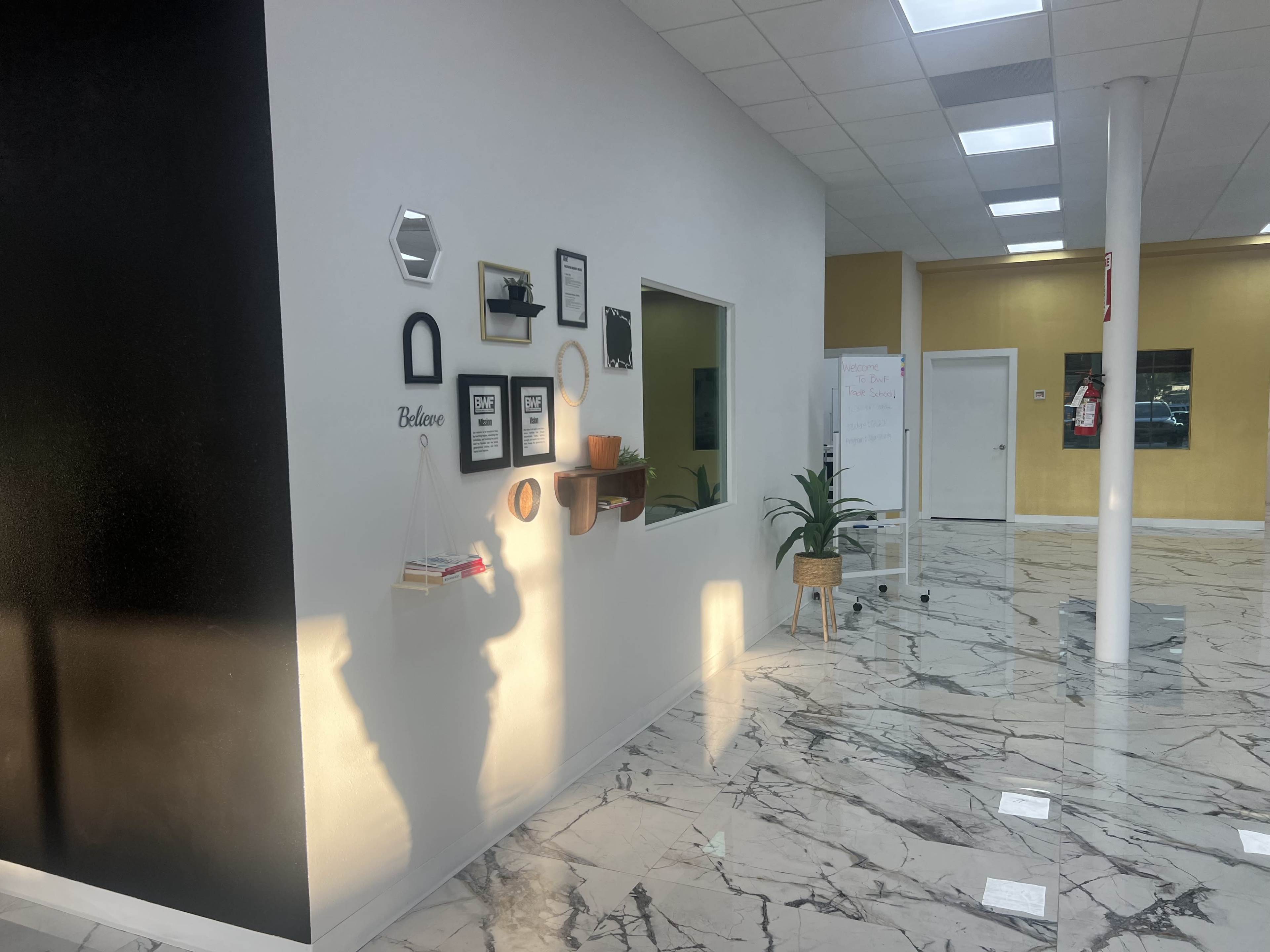 The image shows a well-lit interior space featuring a wall with framed pictures, a decorative shelf, a plant, and marble flooring.