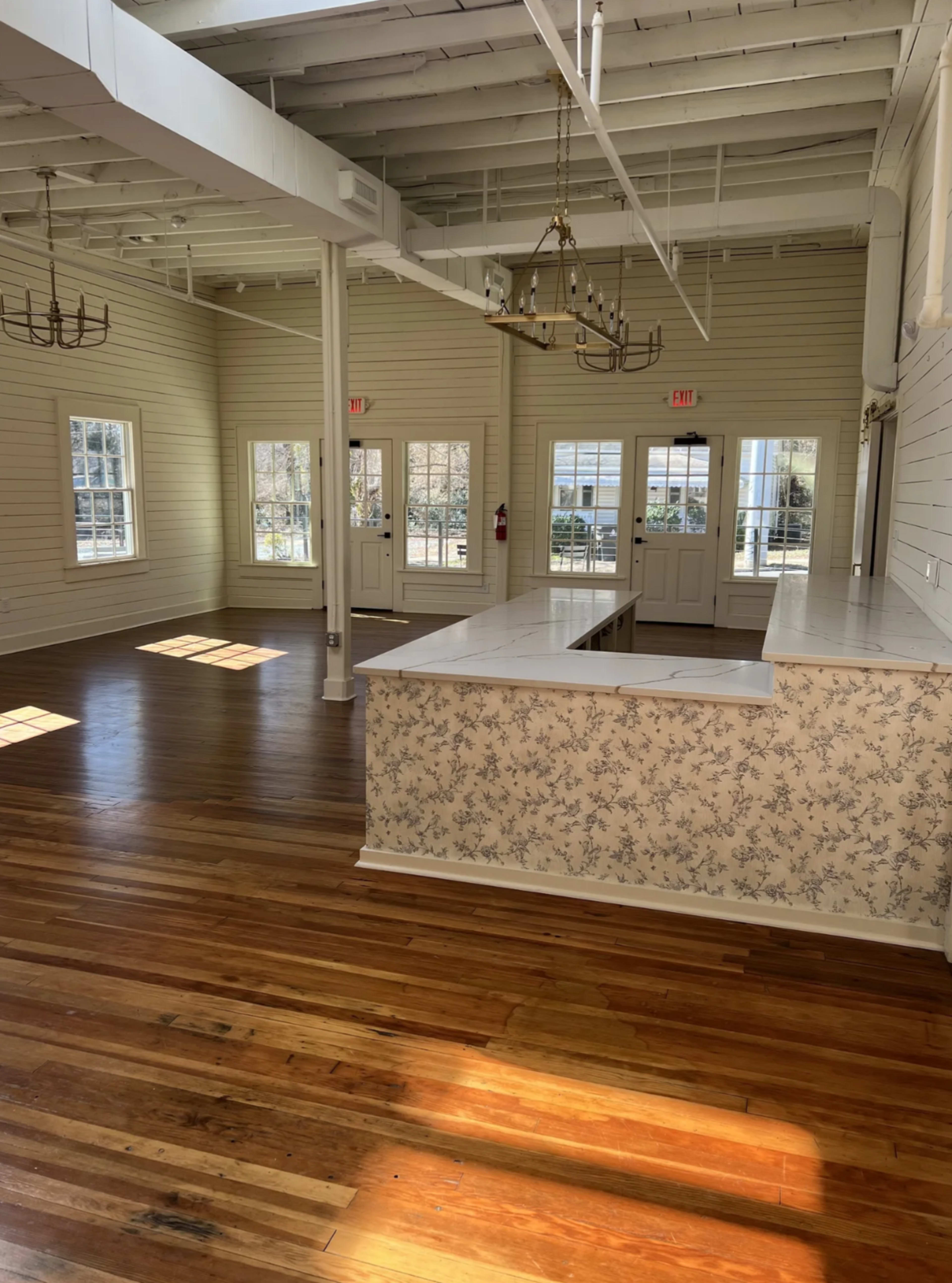 The image shows a bright, spacious interior with wooden floors, large windows, and a countertop against a patterned wall, creating an inviting atmosphere.
