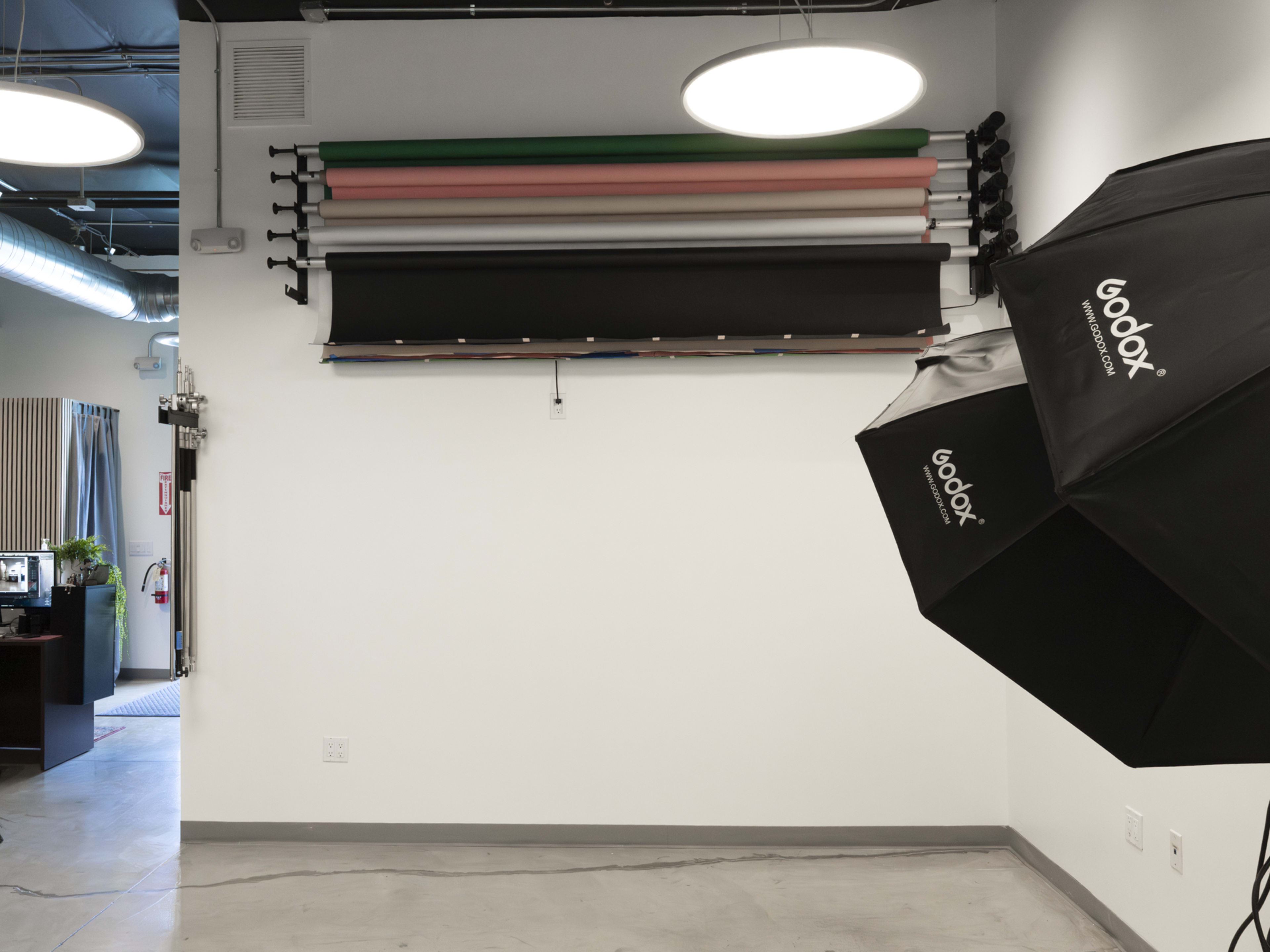 The image shows a blank wall with various rolled backdrops mounted above, and a Godox softbox umbrella positioned to the right.