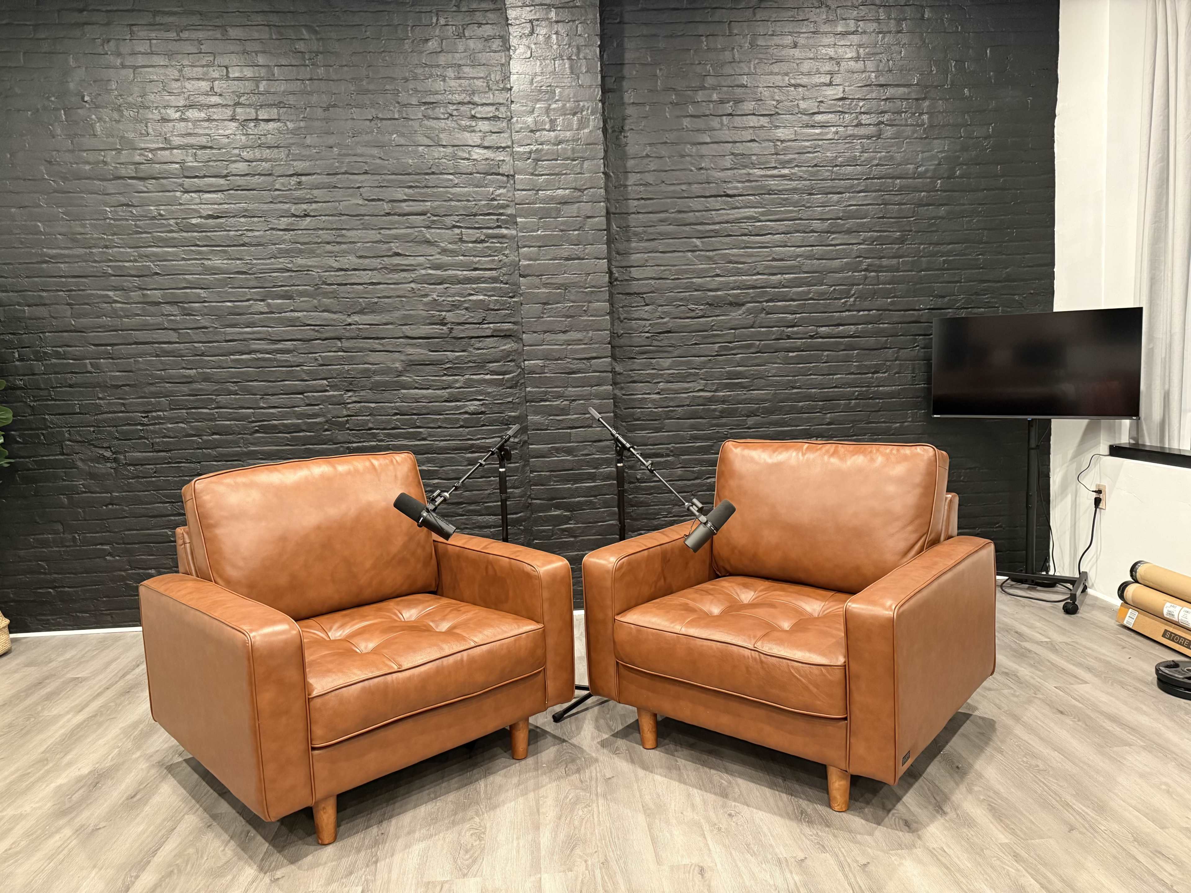 The image shows two brown leather armchairs facing each other in a room with a black brick wall and a television in the background.