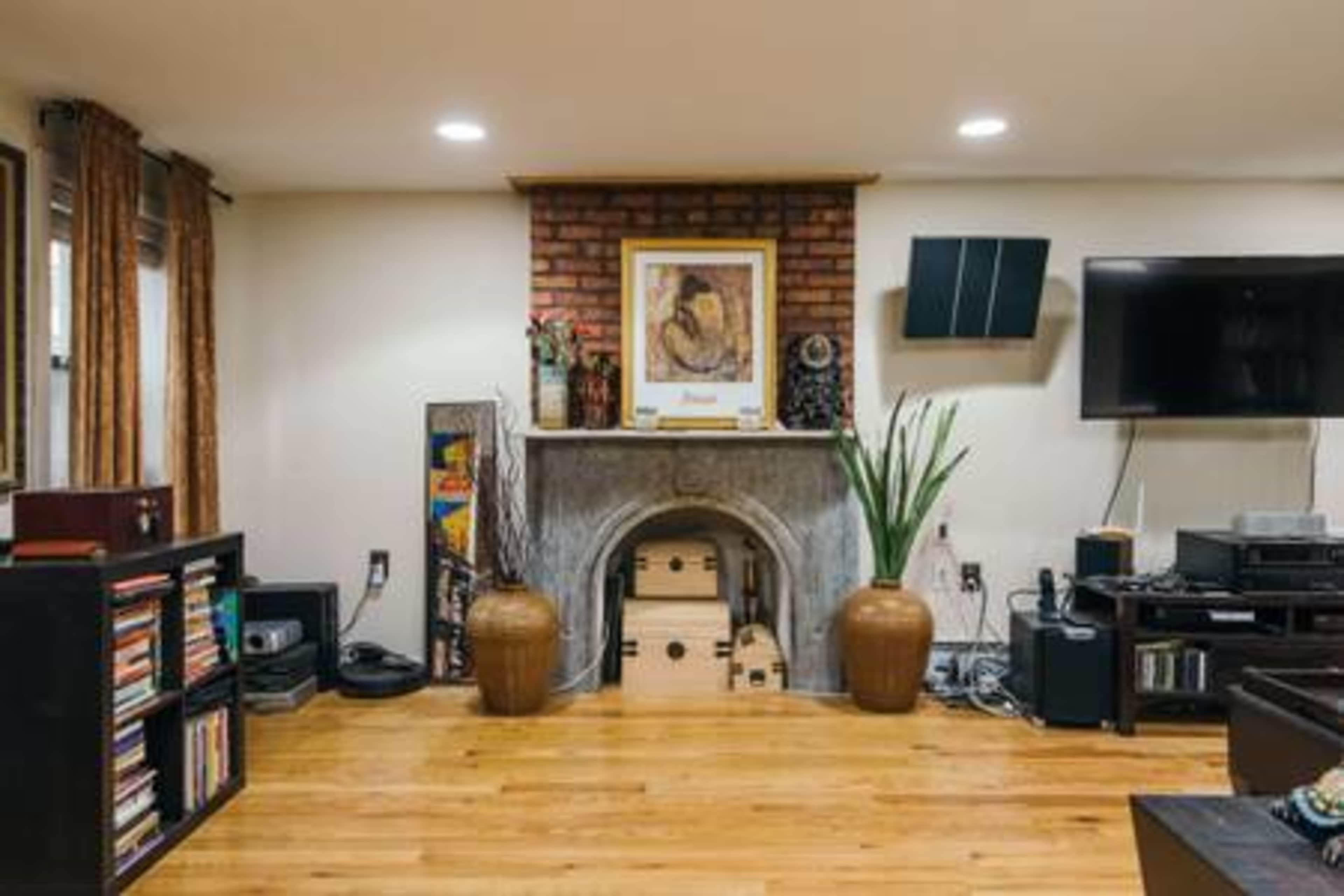 A living room with a brick fireplace, a large framed artwork above it, and a television mounted on the wall, surrounded by shelves with books and various electronics.