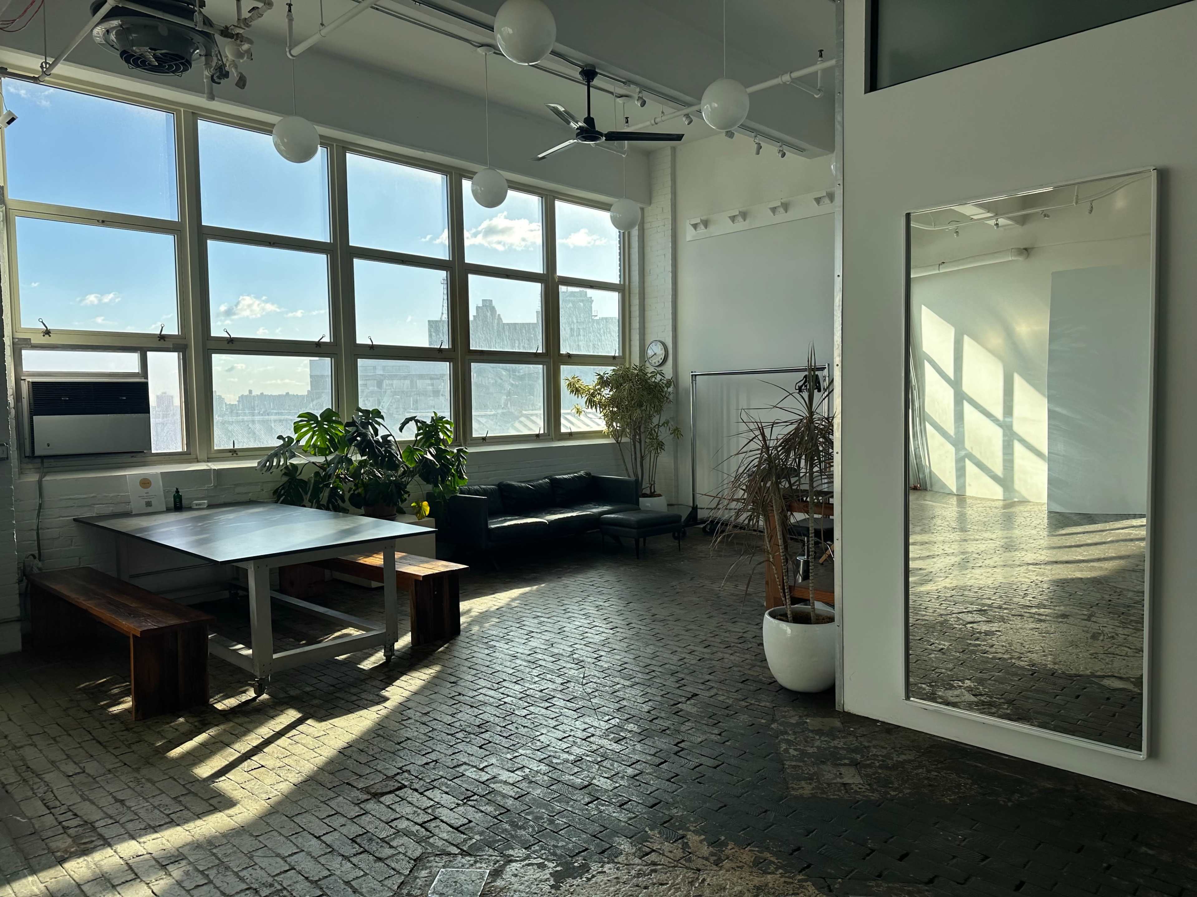 A spacious room with large windows, a dark wooden table and benches, a black sofa, and a tall mirror reflecting light and plants.