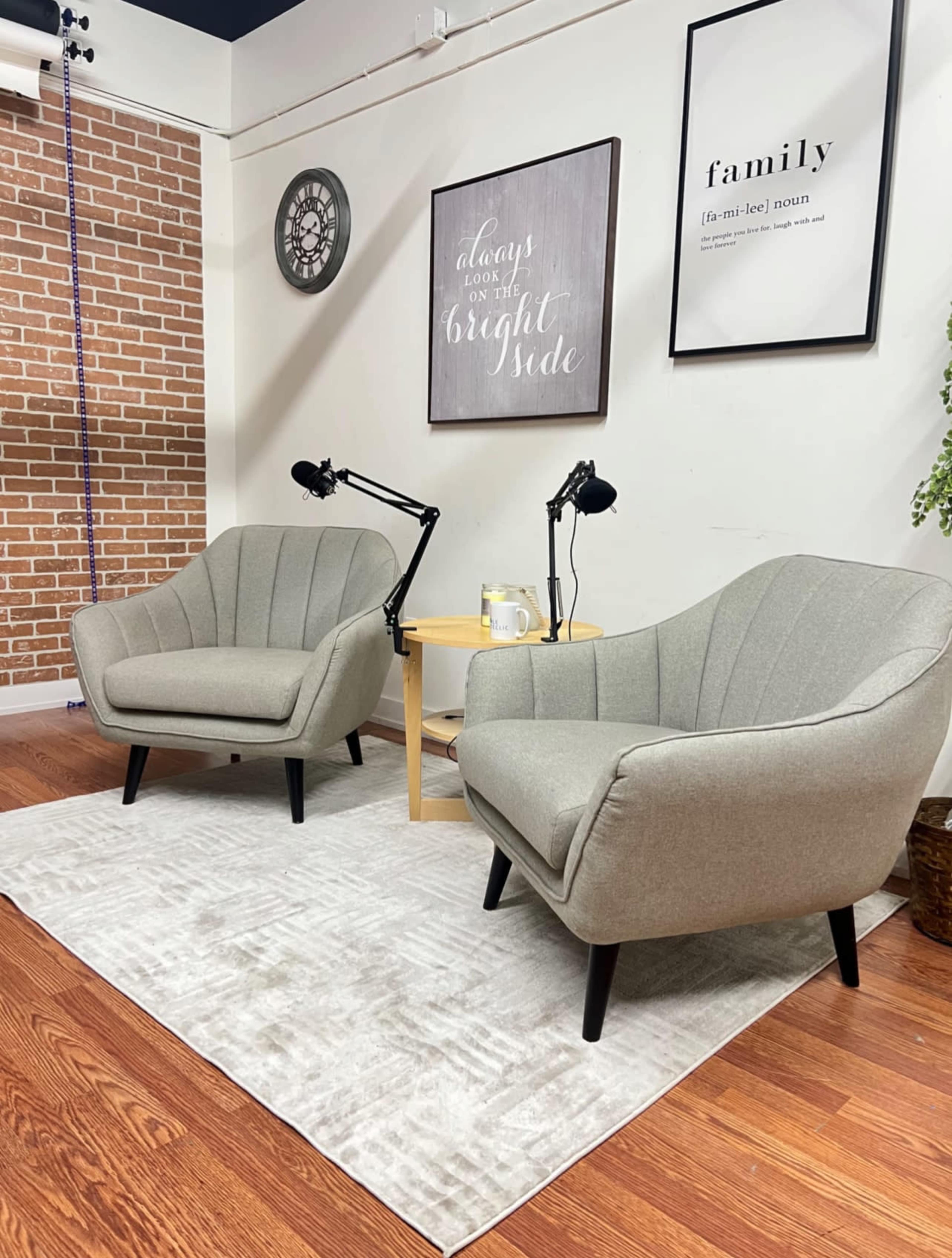 Two gray chairs facing each other on a light-colored rug, with a small round table and microphones positioned nearby in a softly decorated room.