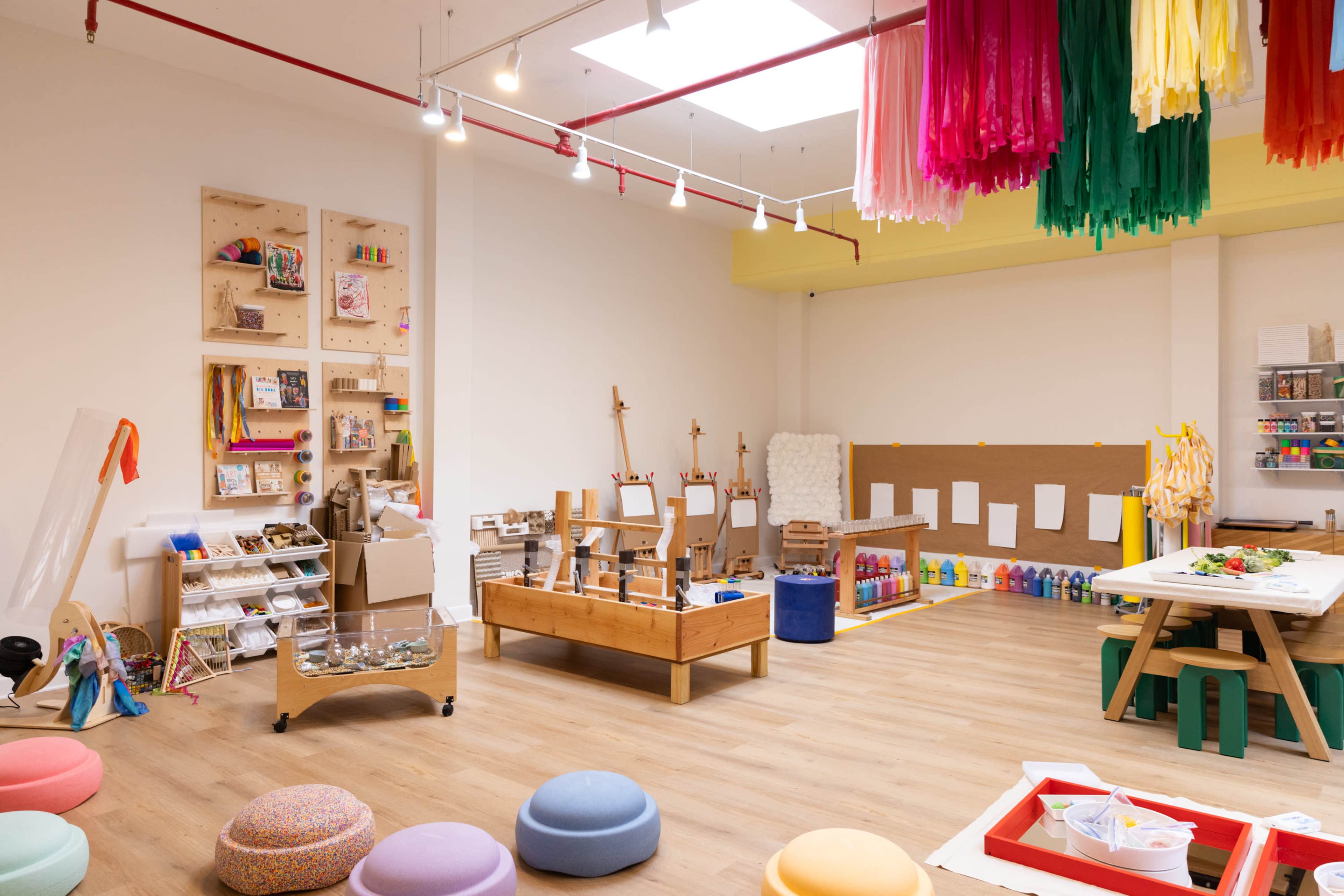 The image shows a brightly lit creative space featuring art supplies, easels, and various colorful seating options.