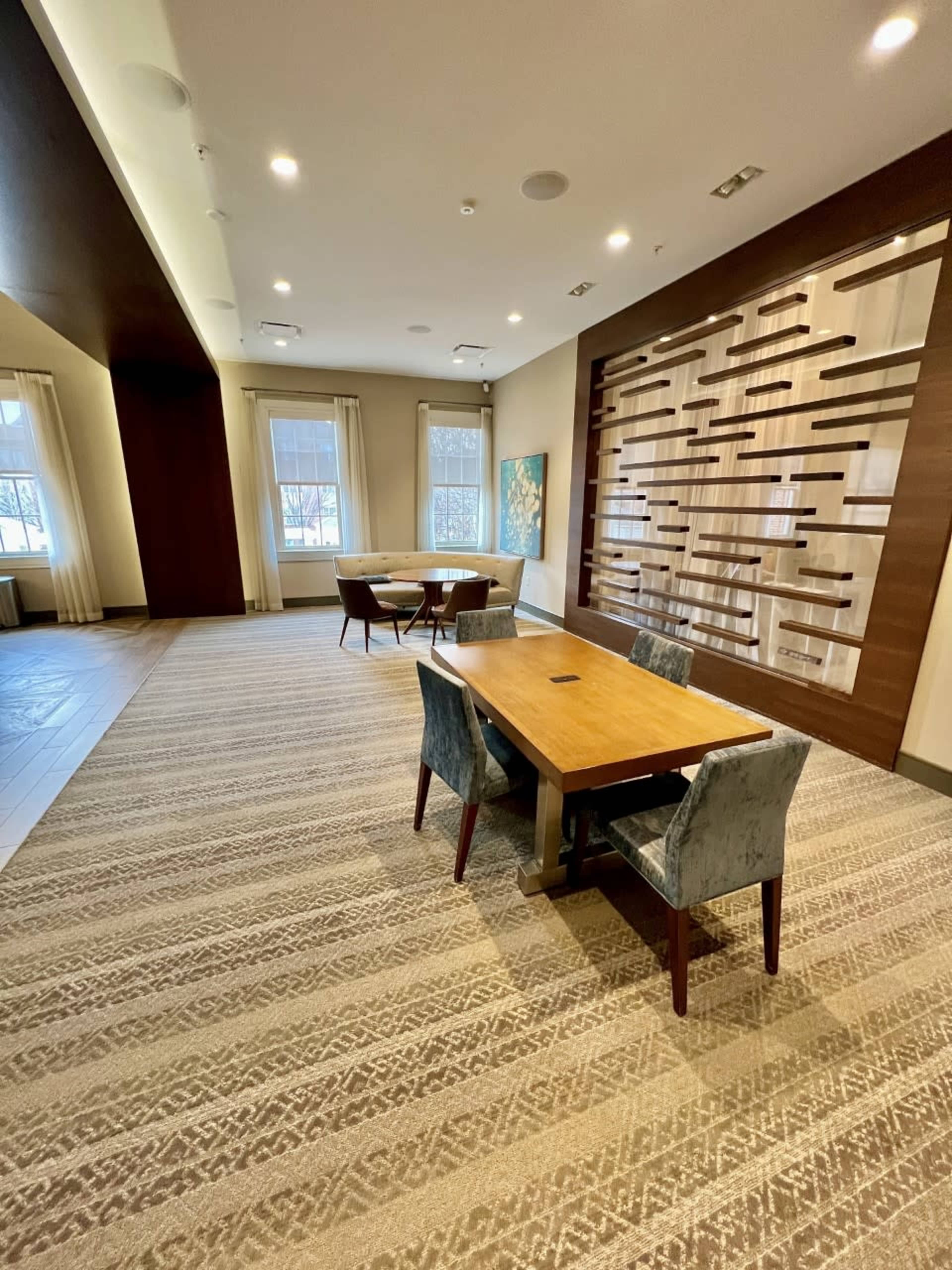 A spacious room with a wooden table and upholstered chairs, featuring large windows and a decorative wall design made of wood panels.