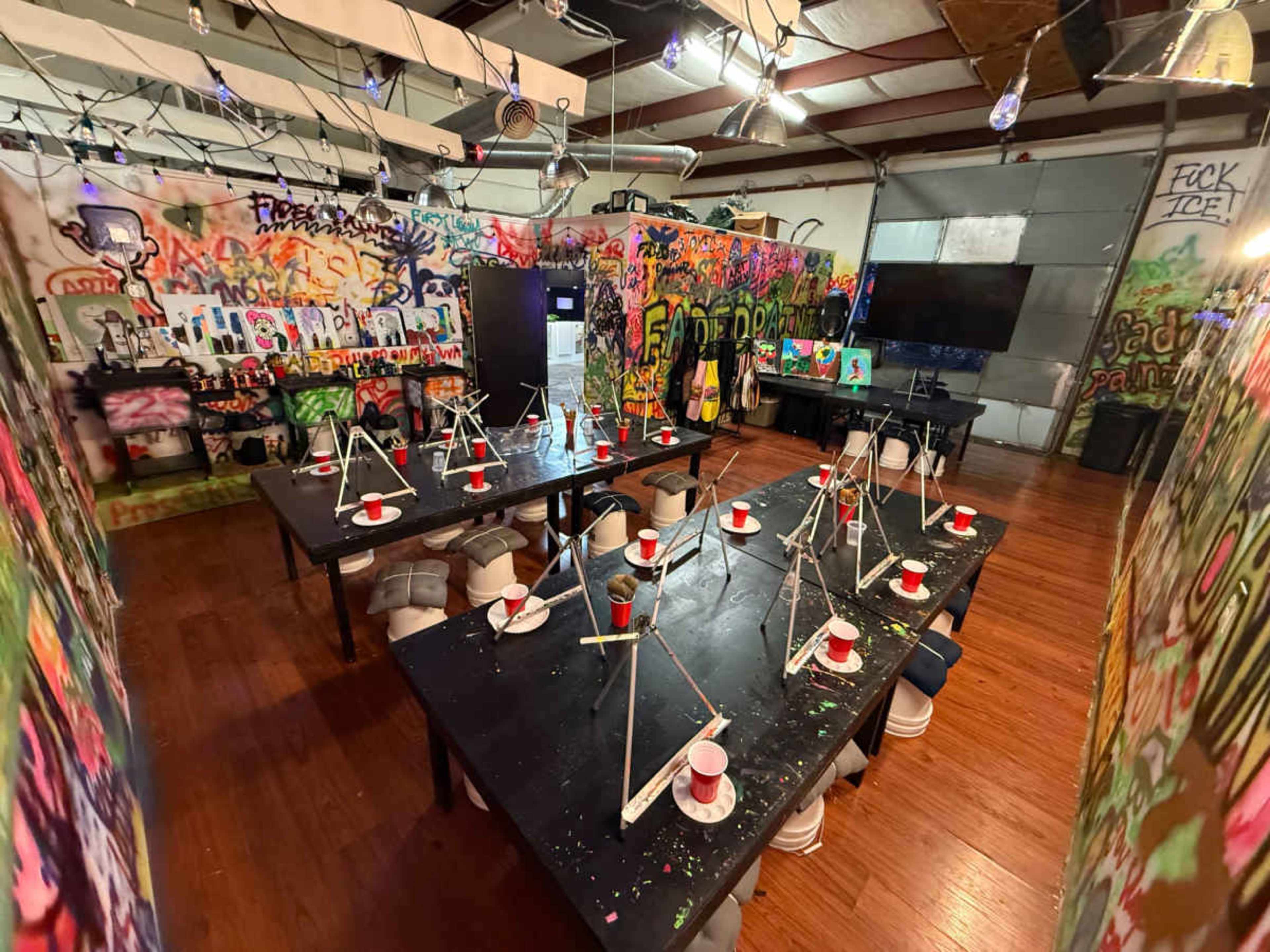 The image shows an art studio with several tables set up for painting, each adorned with paint cups and surrounded by brightly graffiti-covered walls.