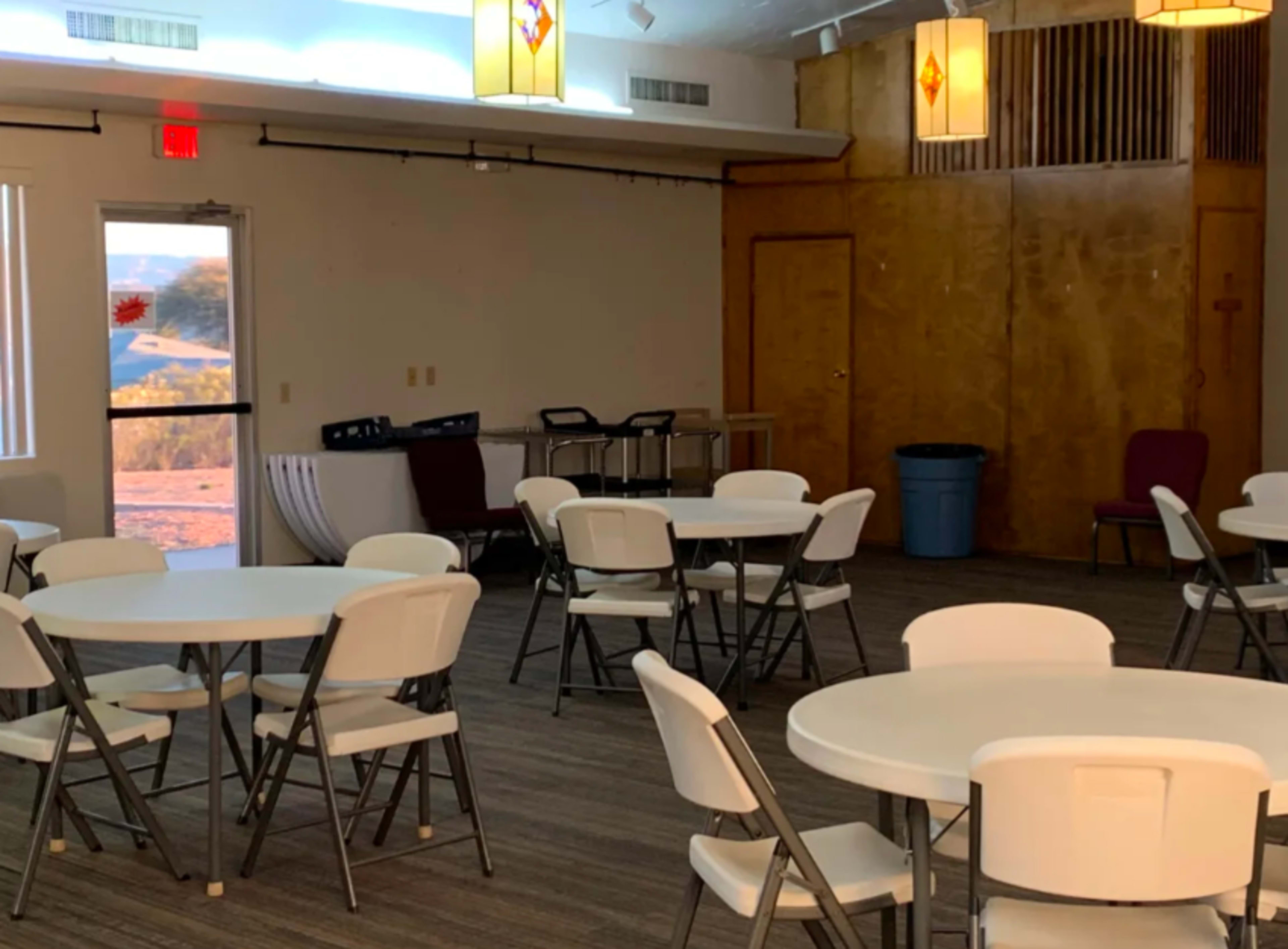 A room set up with several round folding tables and chairs, along with a door leading outside and a garbage bin in the corner.
