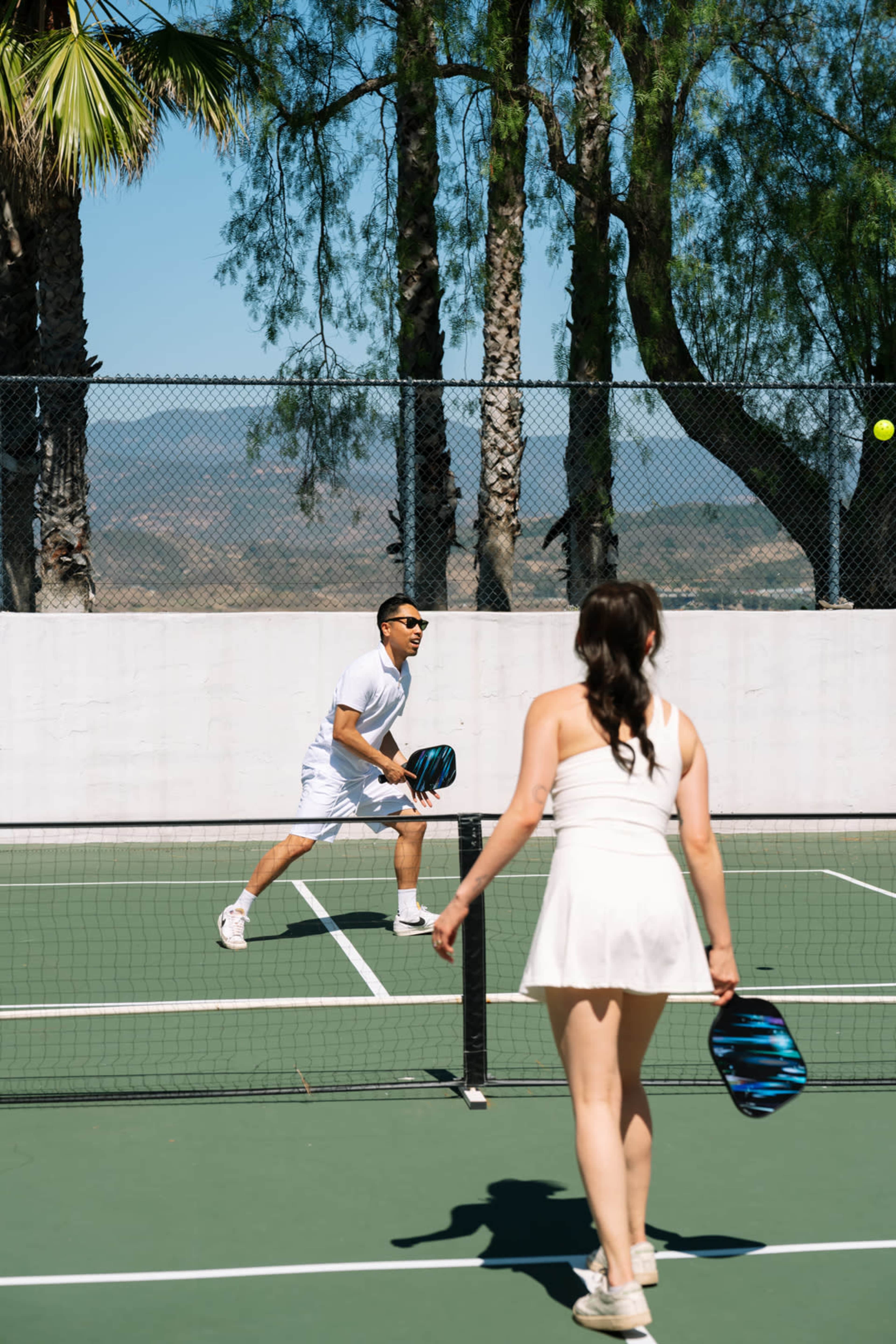 A man and woman play pickleball on an outdoor court surrounded by palm trees, with a ball in mid-air.