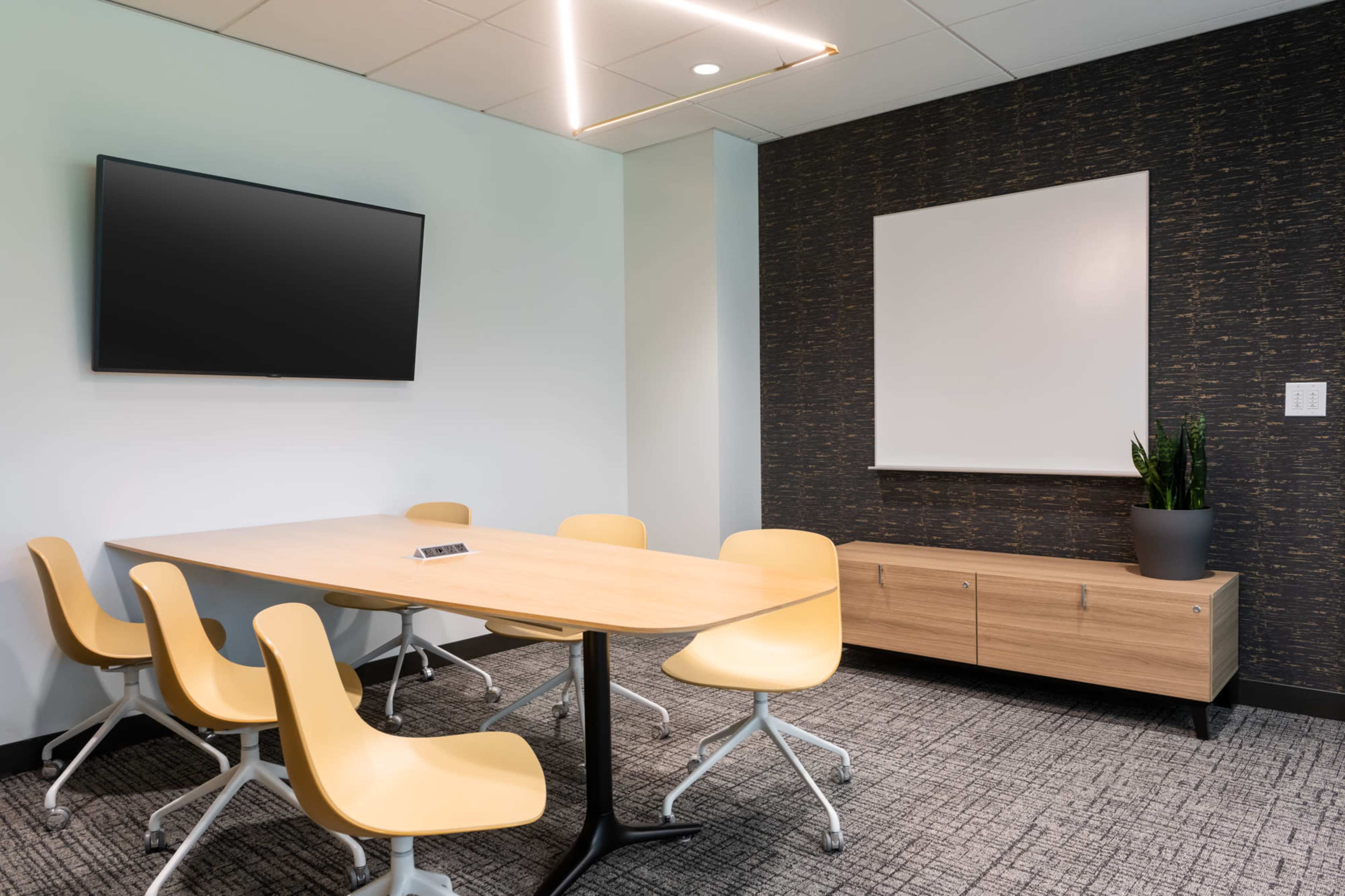 A modern meeting room features a wooden table surrounded by yellow chairs, a wall-mounted television, and a whiteboard on the dark-shaded wall.