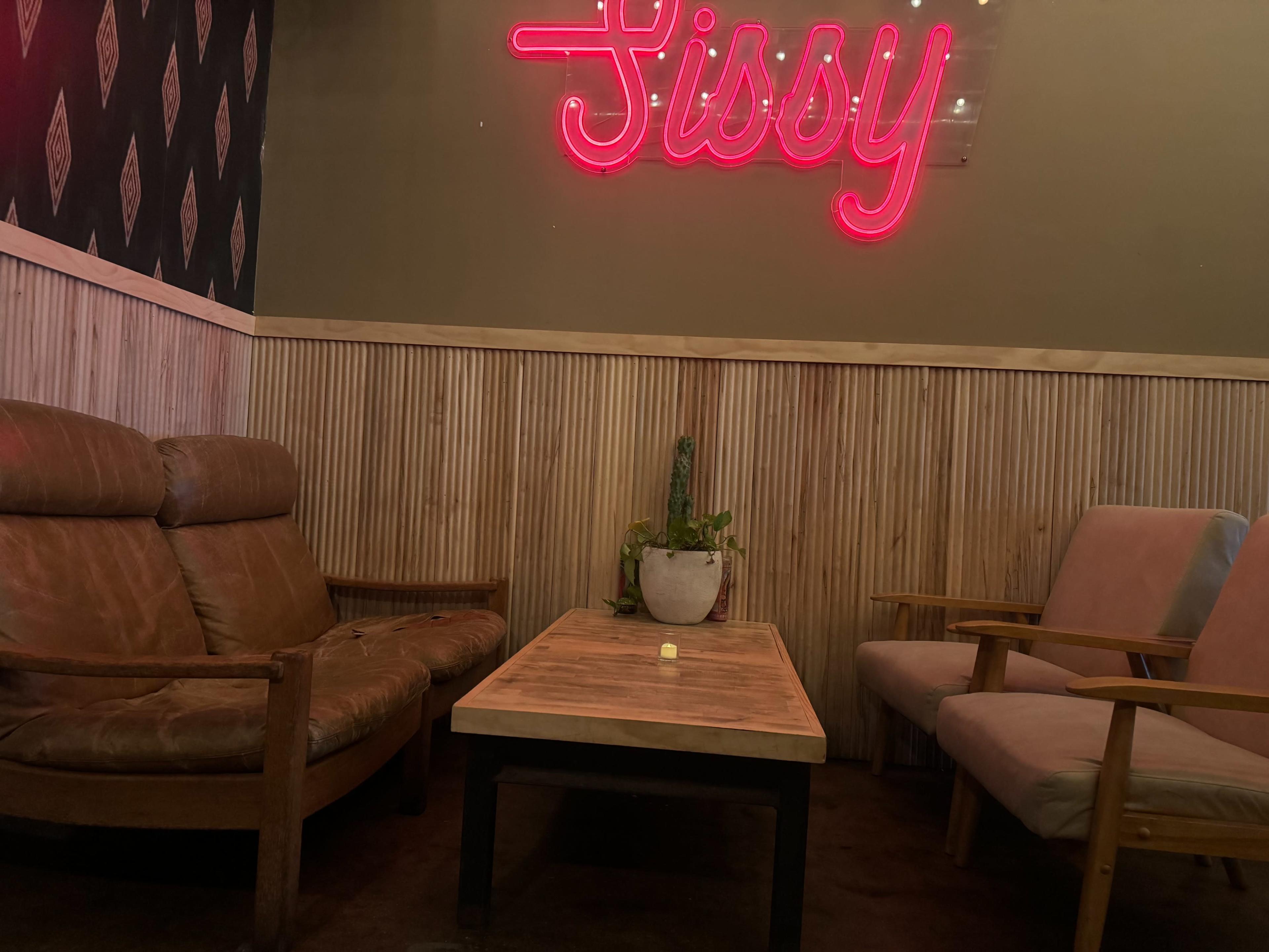 The image shows a cozy seating area with two armchairs, a wooden table, and a neon sign that reads "Sissy" on the wall.