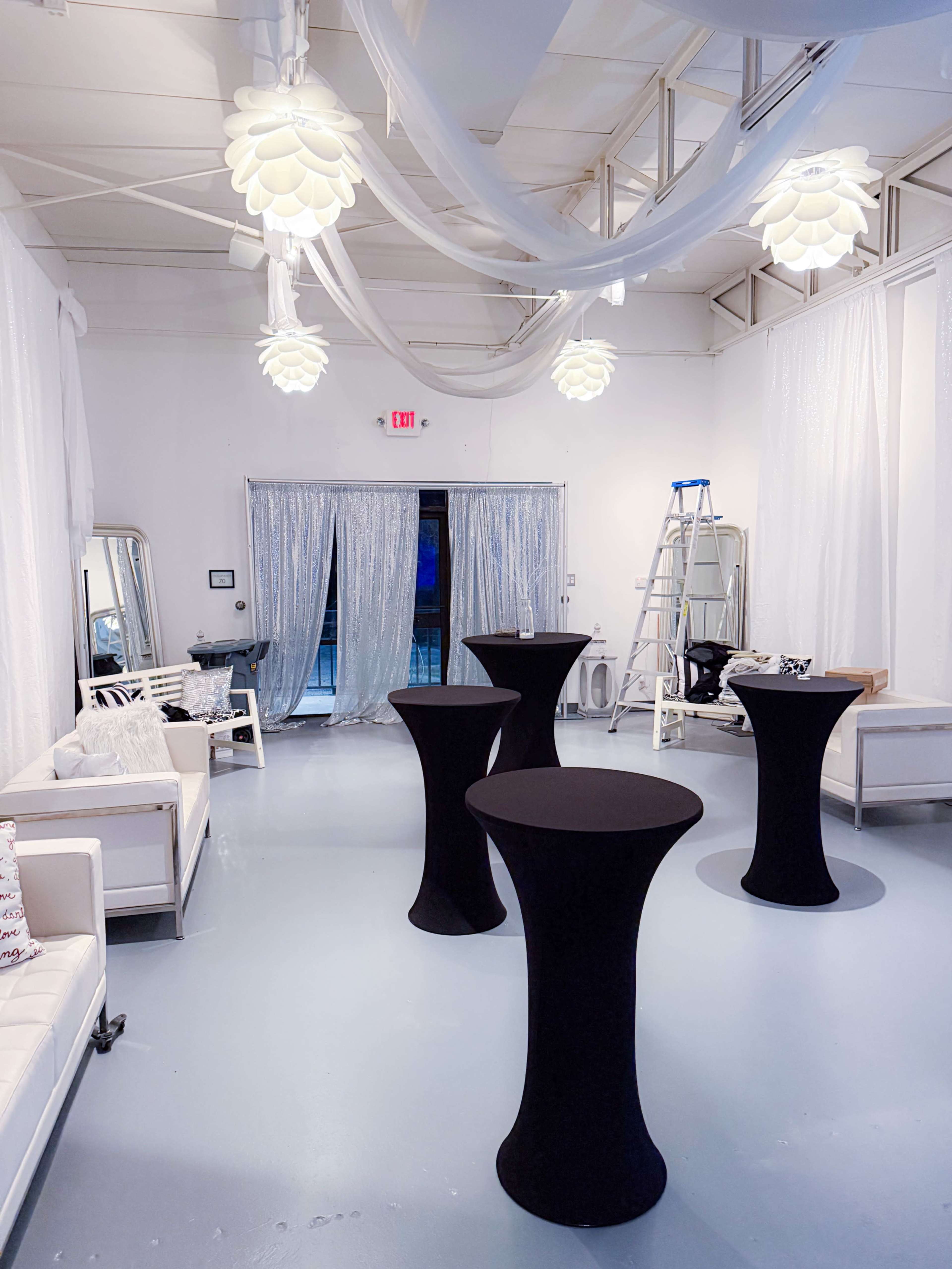 The scene depicts a spacious room with white walls, decorated with draped fabric, featuring several black cocktail tables and illuminated by pendant lights.