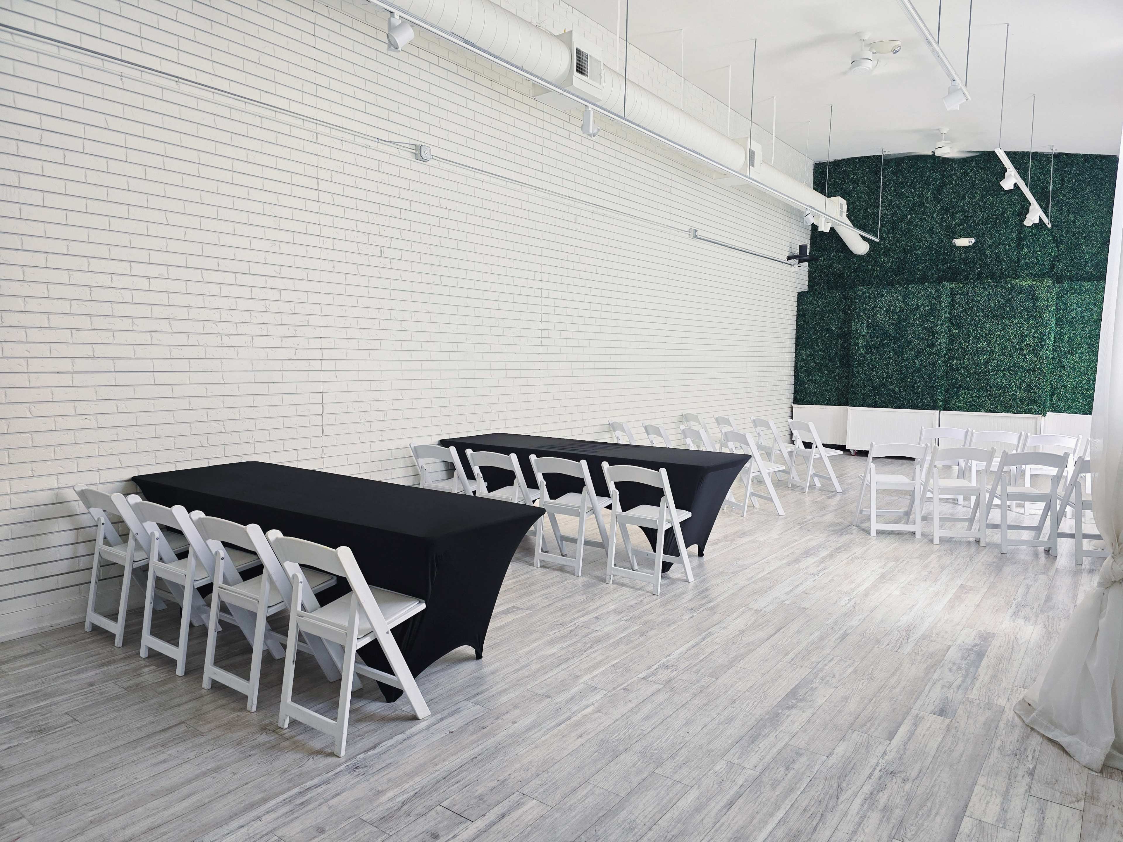 A spacious room with white walls, featuring two black tablecloth-covered tables and several white folding chairs arranged neatly.