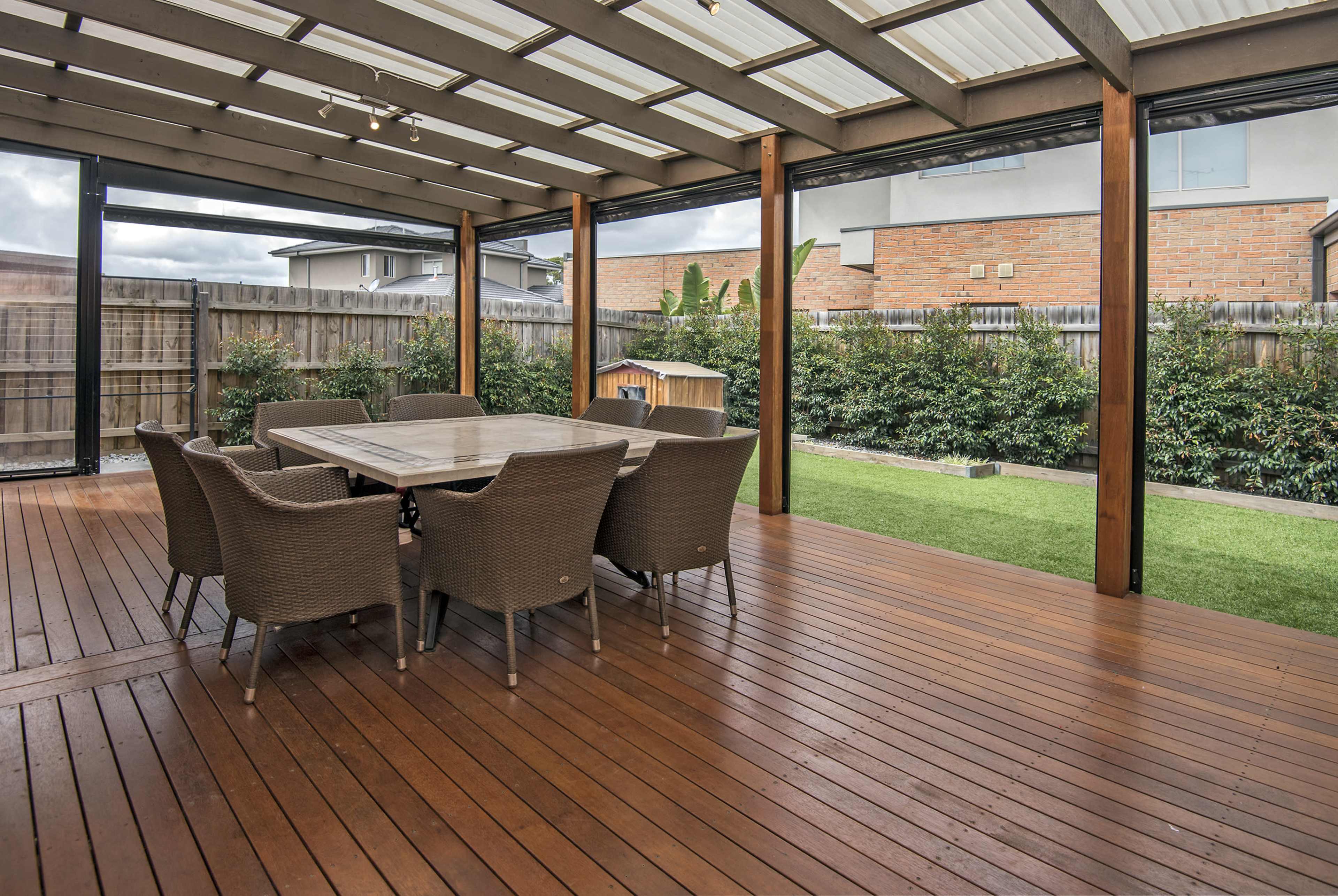 A covered outdoor patio with a wooden deck, featuring a large dining table surrounded by wicker chairs, overlooking a grassy yard and shrubs.
