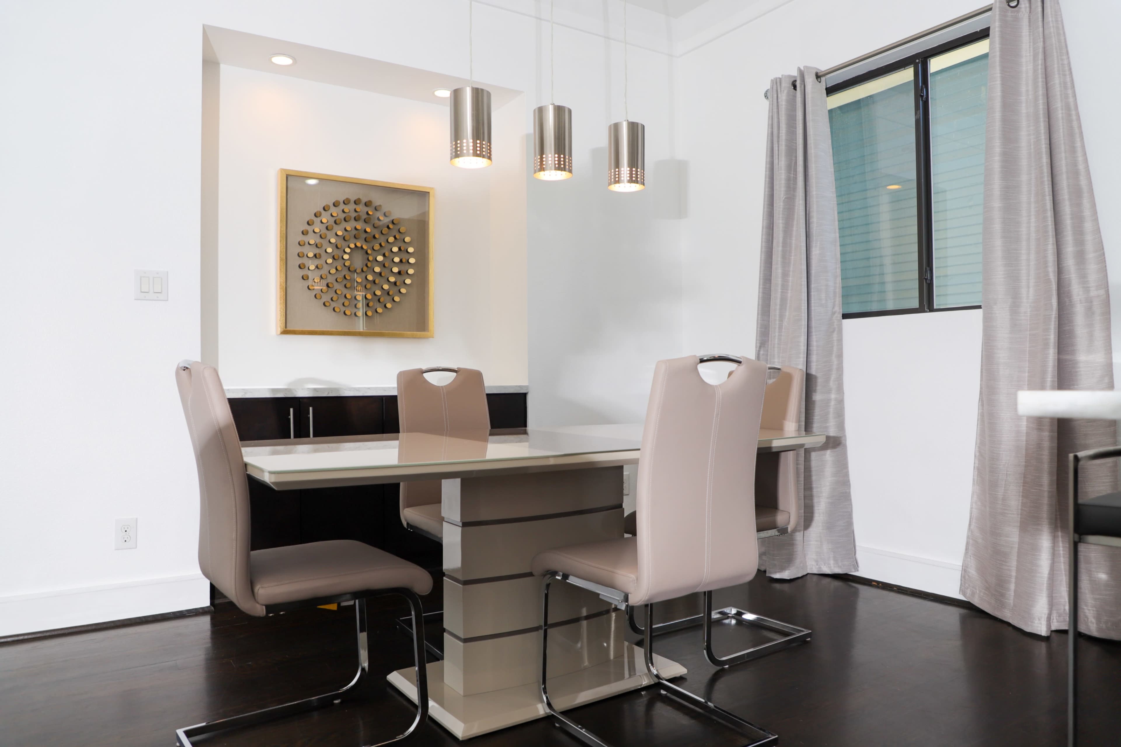 A modern dining area featuring a rectangular table with four upholstered chairs, situated under three pendant lights, alongside a wall with a decorative circular wall art.
