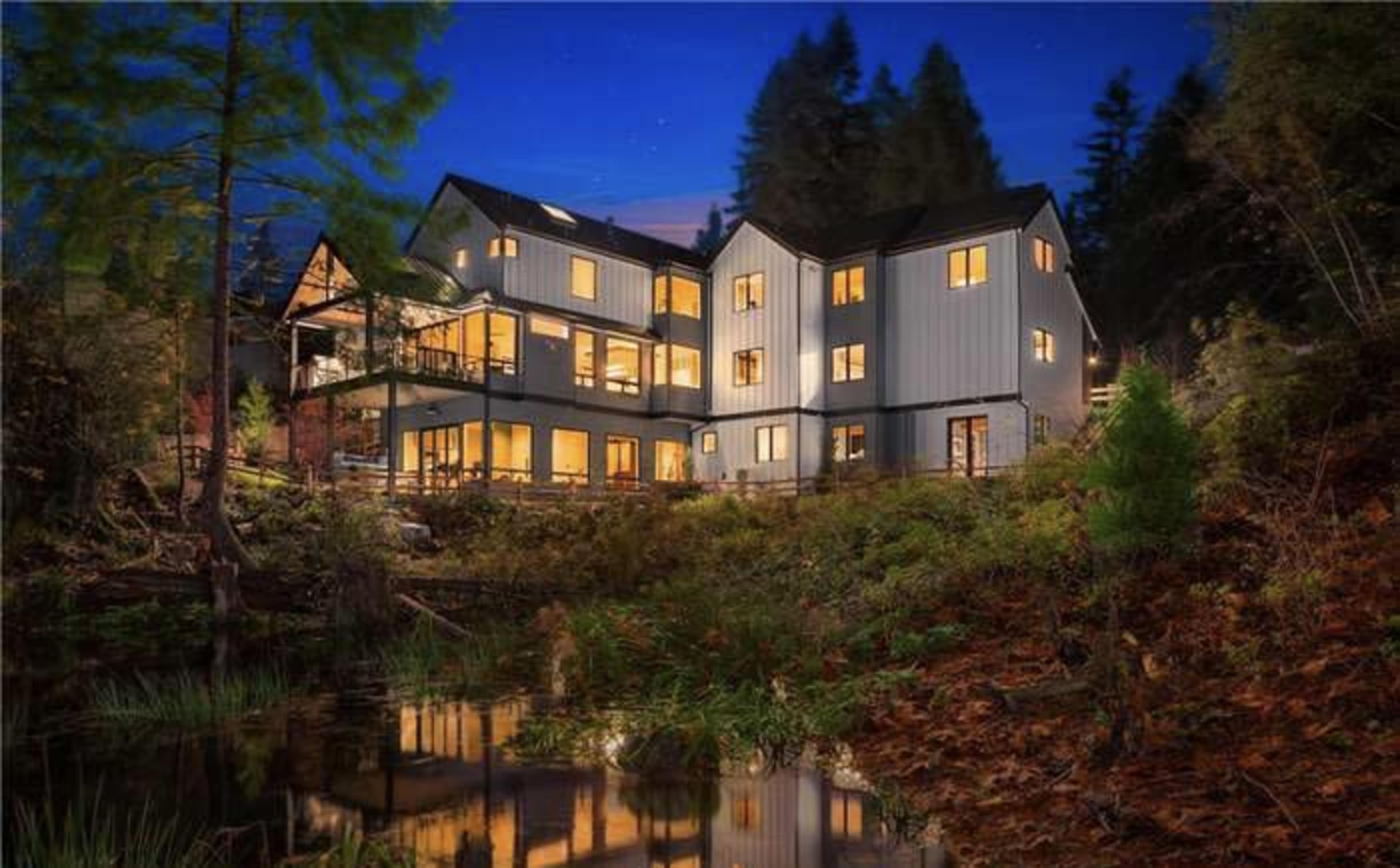 A modern house with multiple levels and large windows is illuminated at dusk, situated next to a tranquil body of water and surrounded by trees.