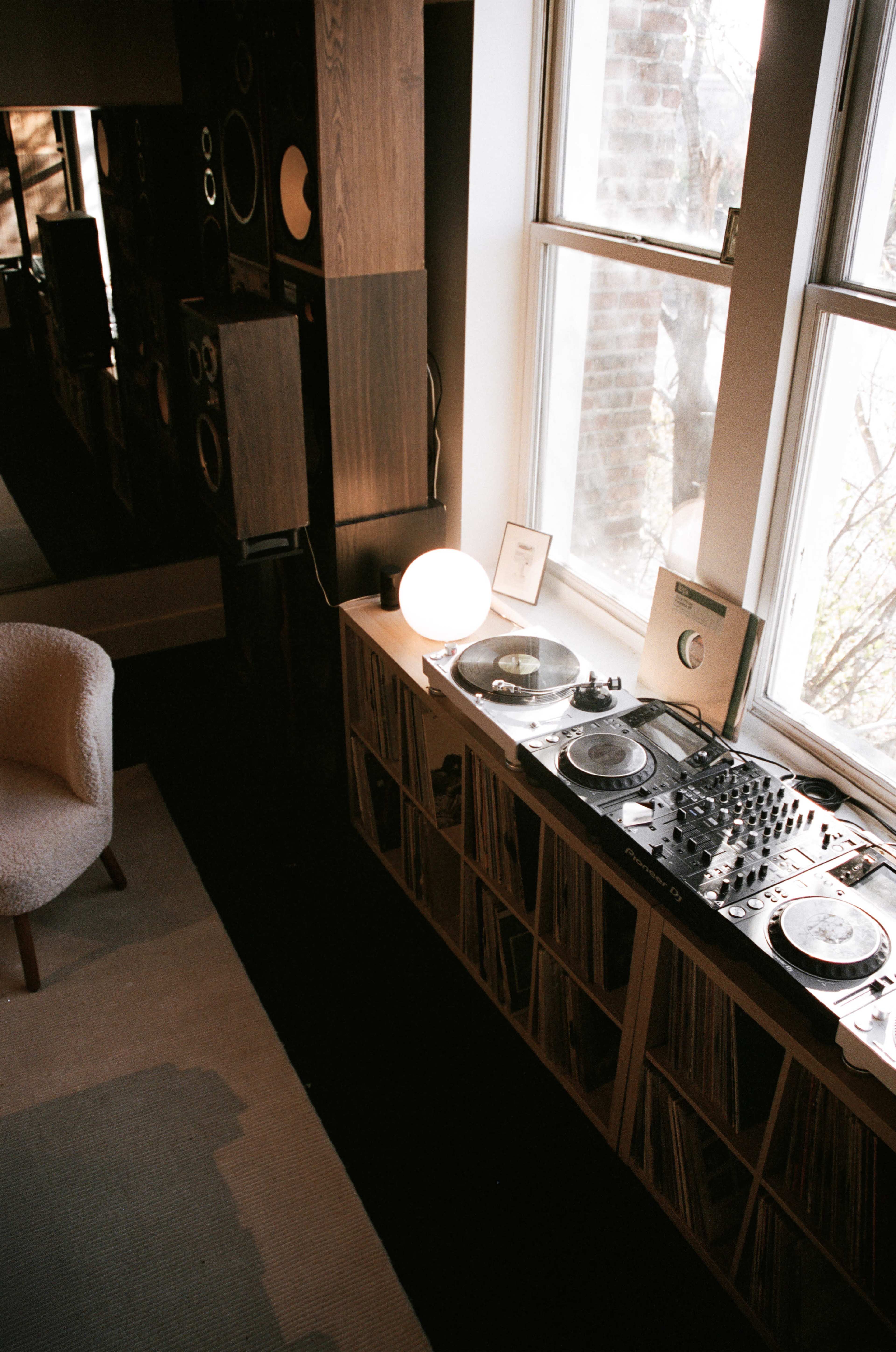 The image shows a cozy interior with a DJ turntable setup, vinyl records stored on a shelf, a round lamp, and a chair positioned nearby.