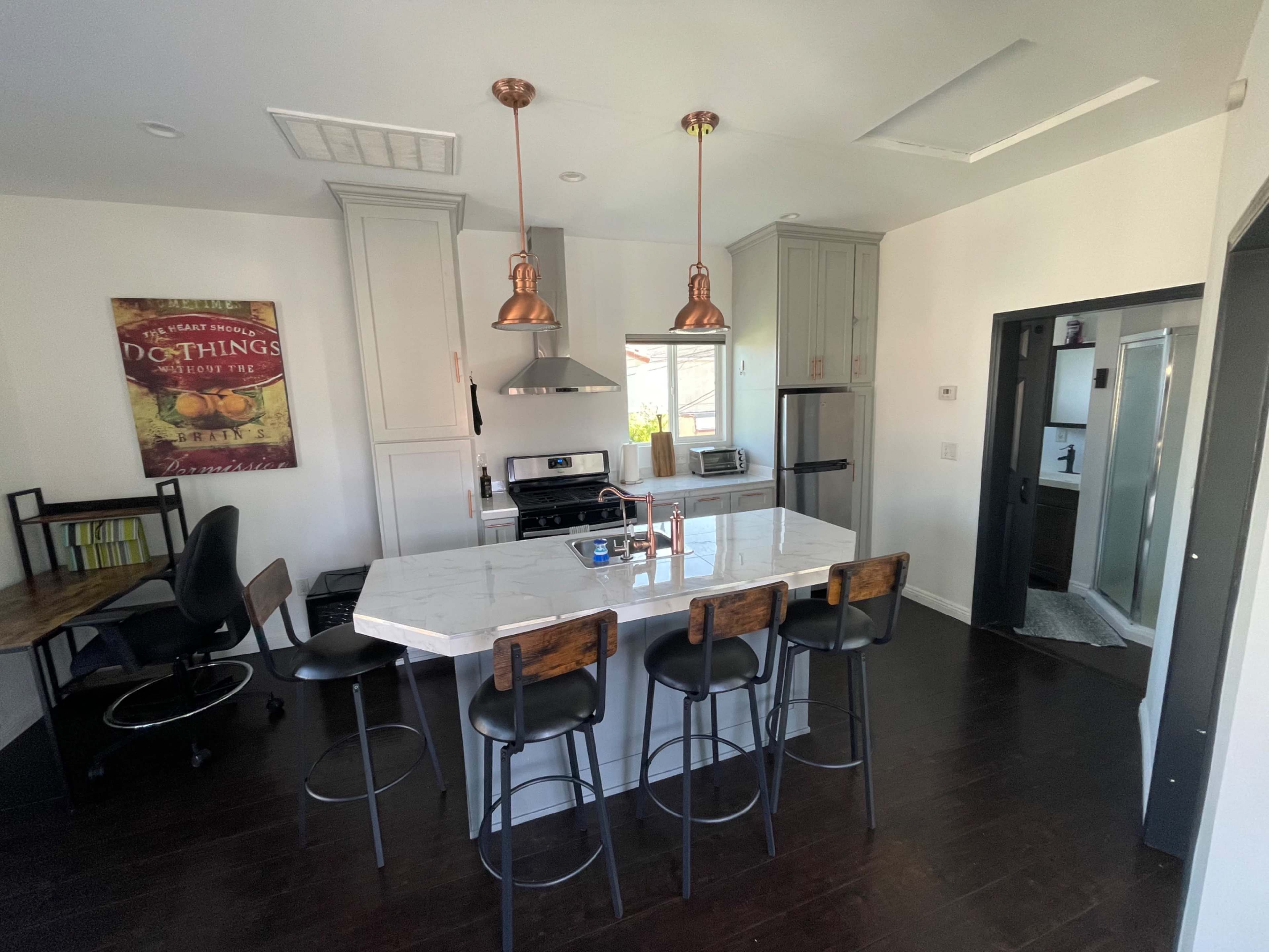 The image shows a modern kitchen featuring a central island with four black bar stools, stainless steel appliances, and copper pendant lights hanging above.