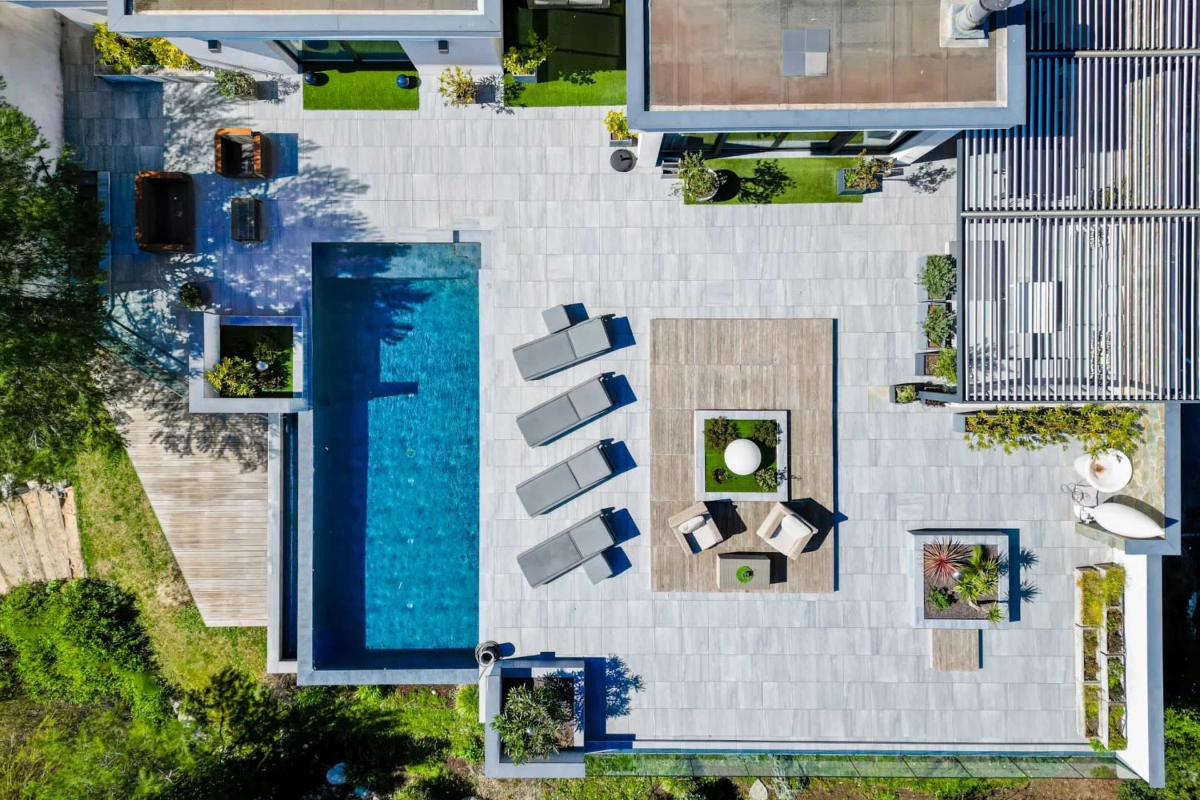 The image shows an aerial view of a modern outdoor patio featuring a rectangular swimming pool, lounge chairs, and a seating area with a table and chairs surrounded by greenery.