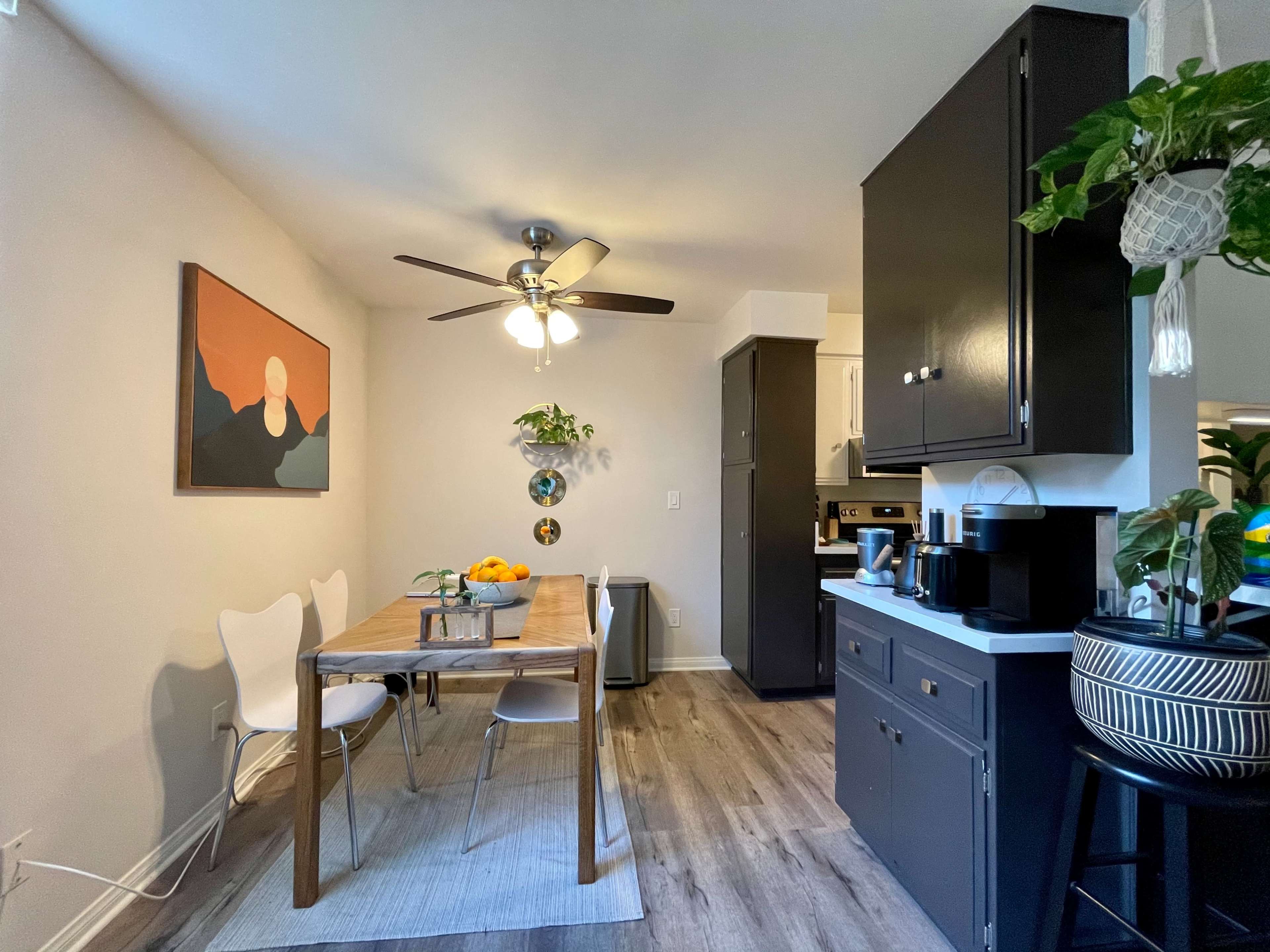 A modern kitchen features a wooden dining table with chairs, a ceiling fan, and black cabinetry.