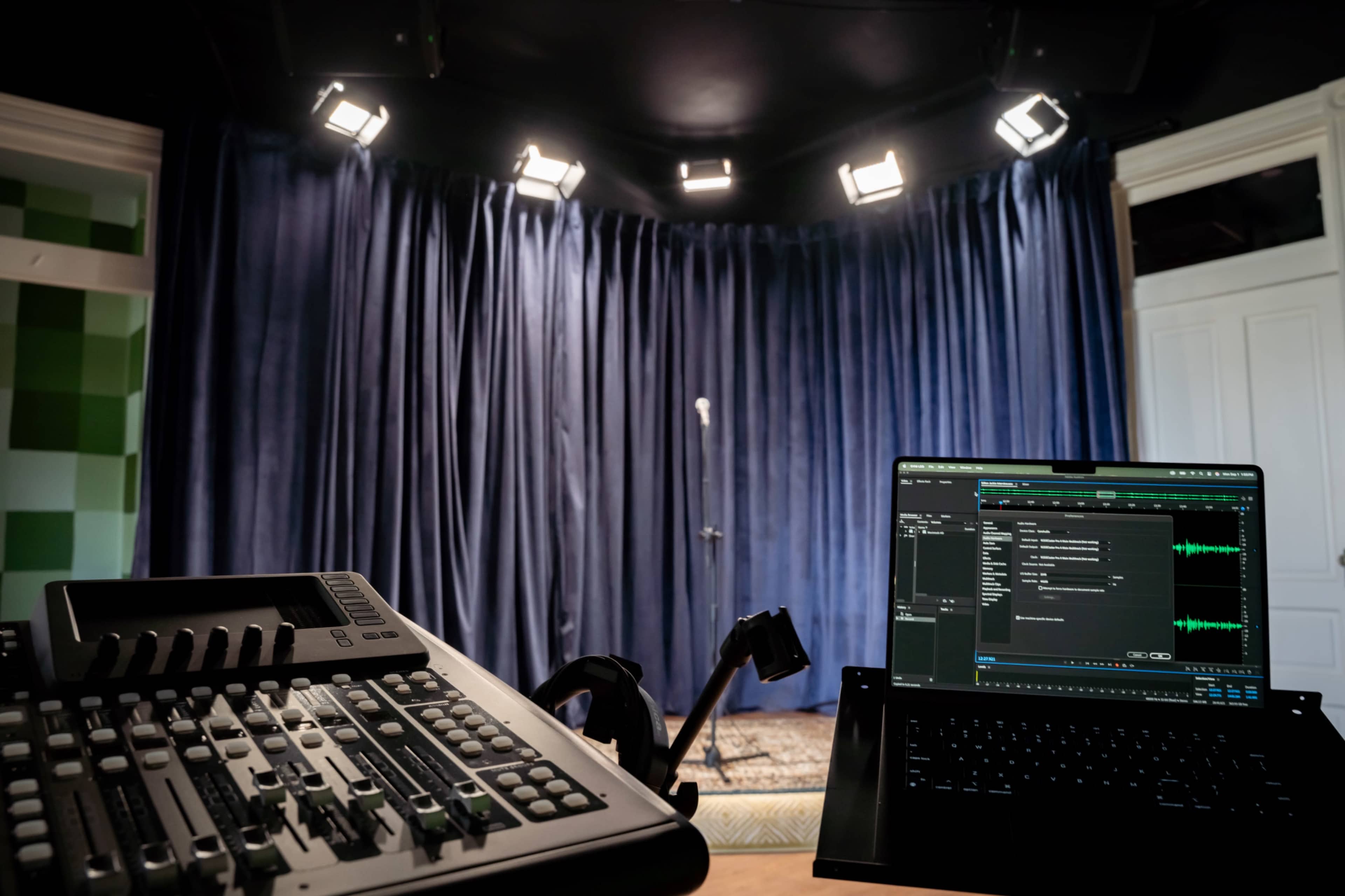 The image shows a sound mixing console and a laptop in the foreground, with a microphone and a curtain backdrop set for a performance in the background.