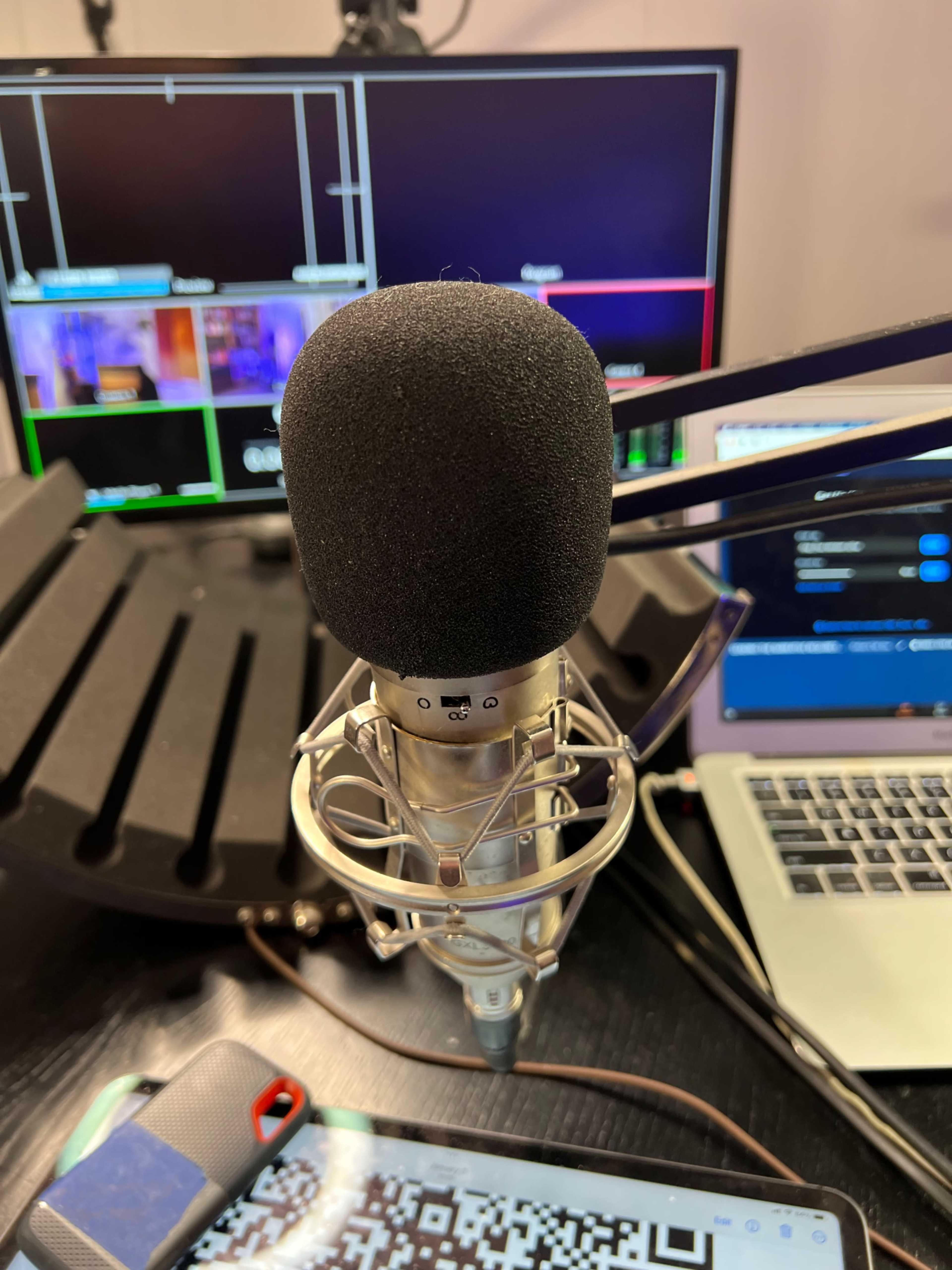 A microphone is positioned on a table with multiple screens and a laptop in the background.