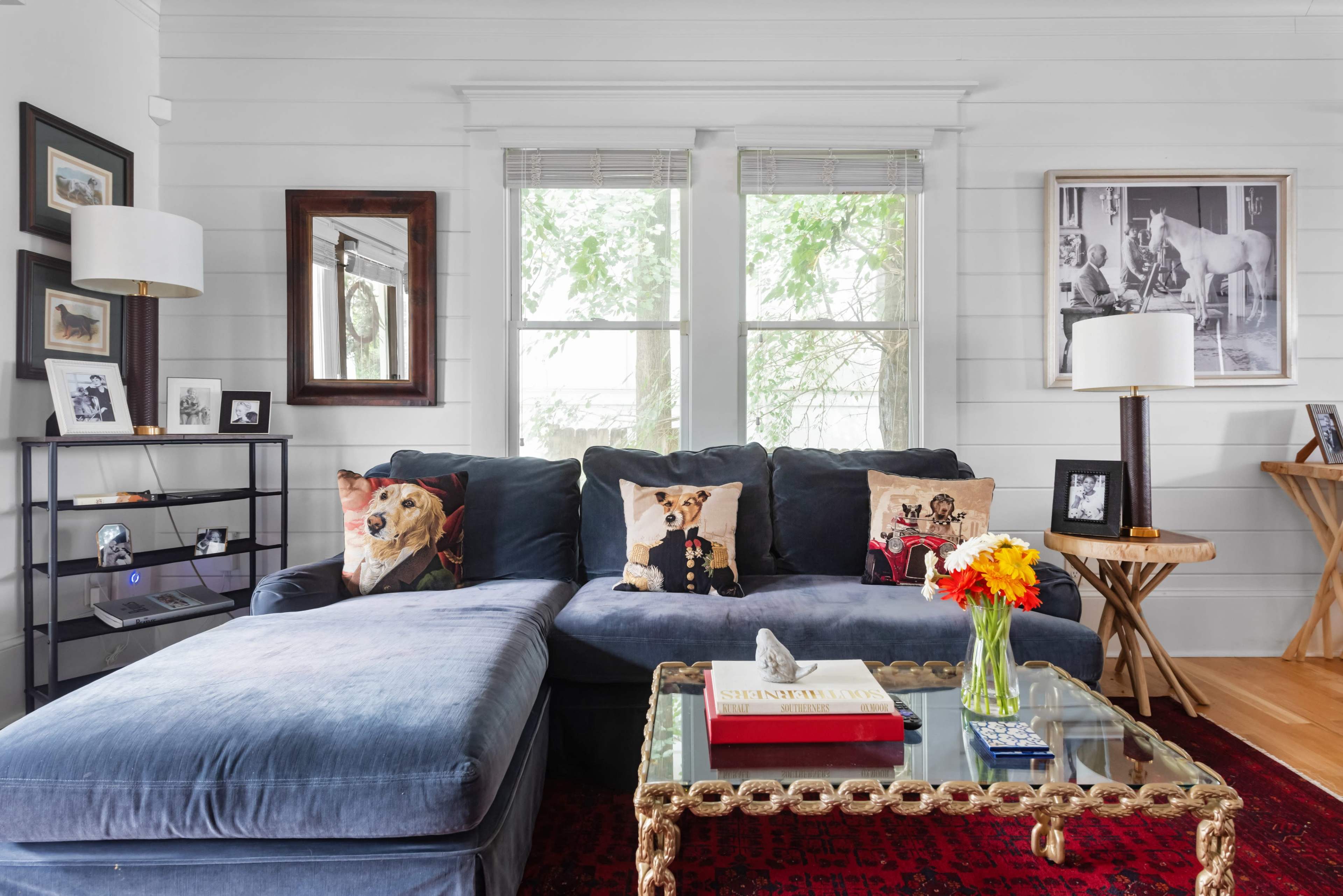 The living room features a blue sectional sofa adorned with decorative pillows, a glass coffee table, and various framed artworks on the white walls.