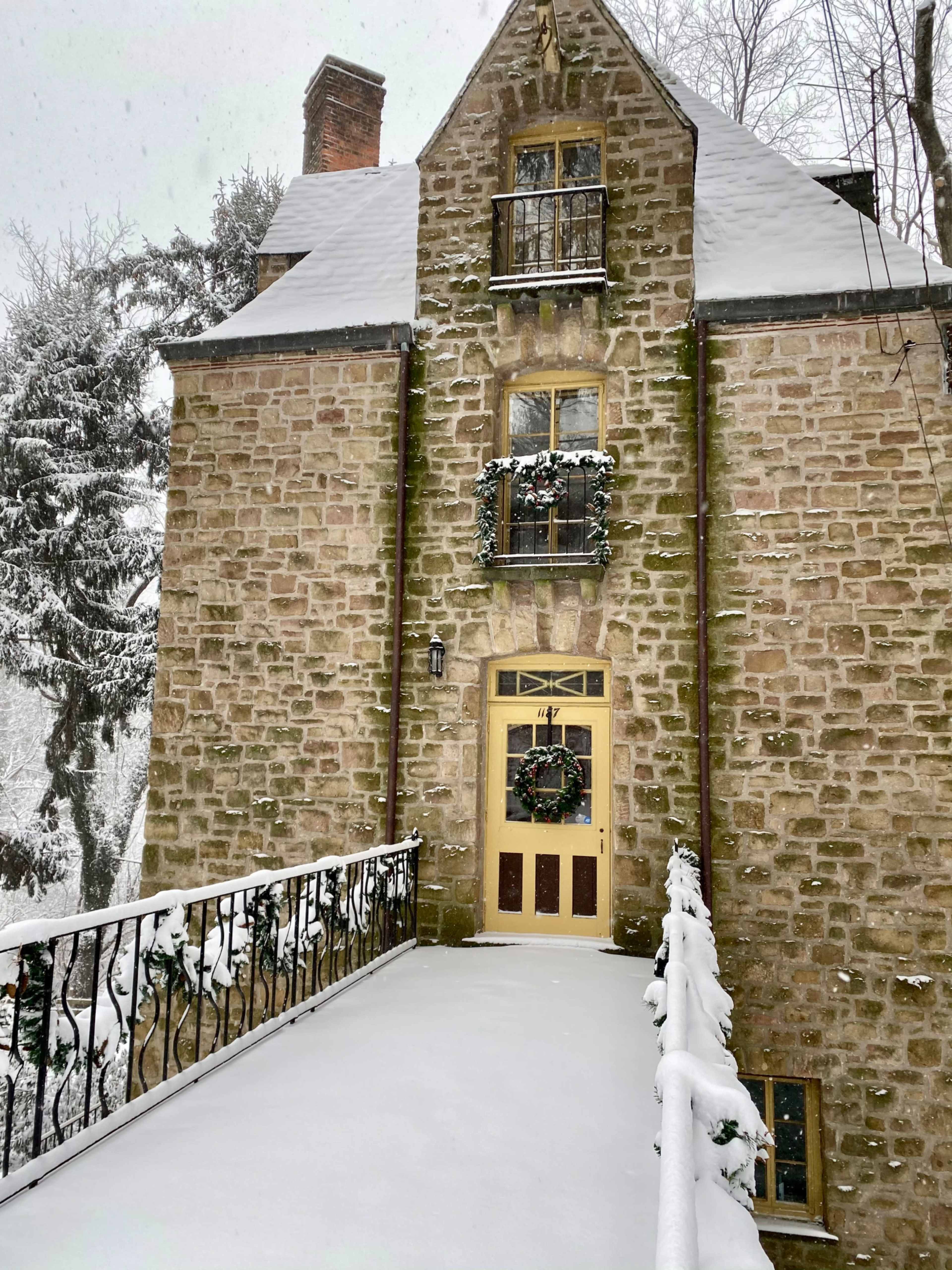 A stone house with a front door and decorated windows is covered in snow, with a snow-laden pathway leading to the entrance.