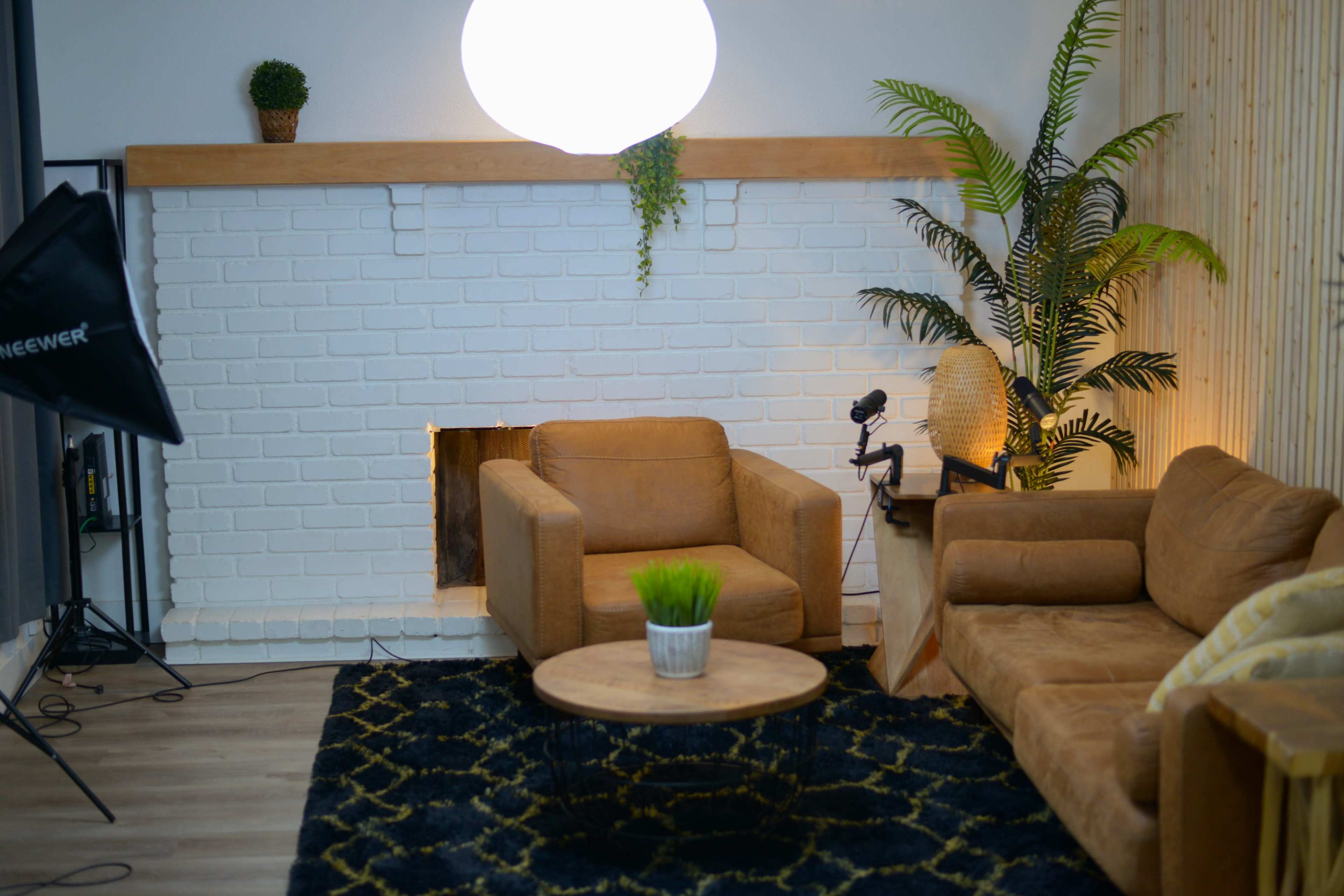 A cozy living room with a white brick fireplace, two brown armchairs, a round coffee table with a plant, and a large light fixture above.