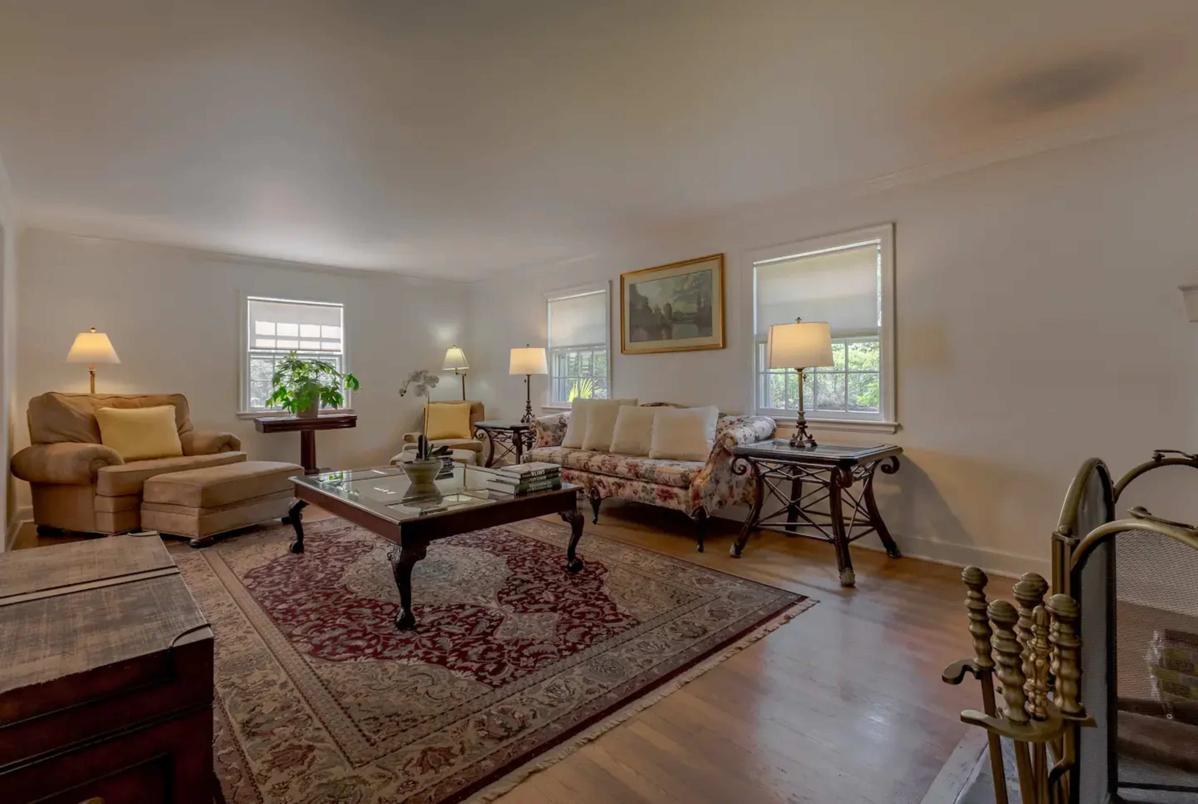 A living room featuring a floral-patterned couch, two armchairs, a wooden coffee table, and a rug, with natural light coming through two windows.