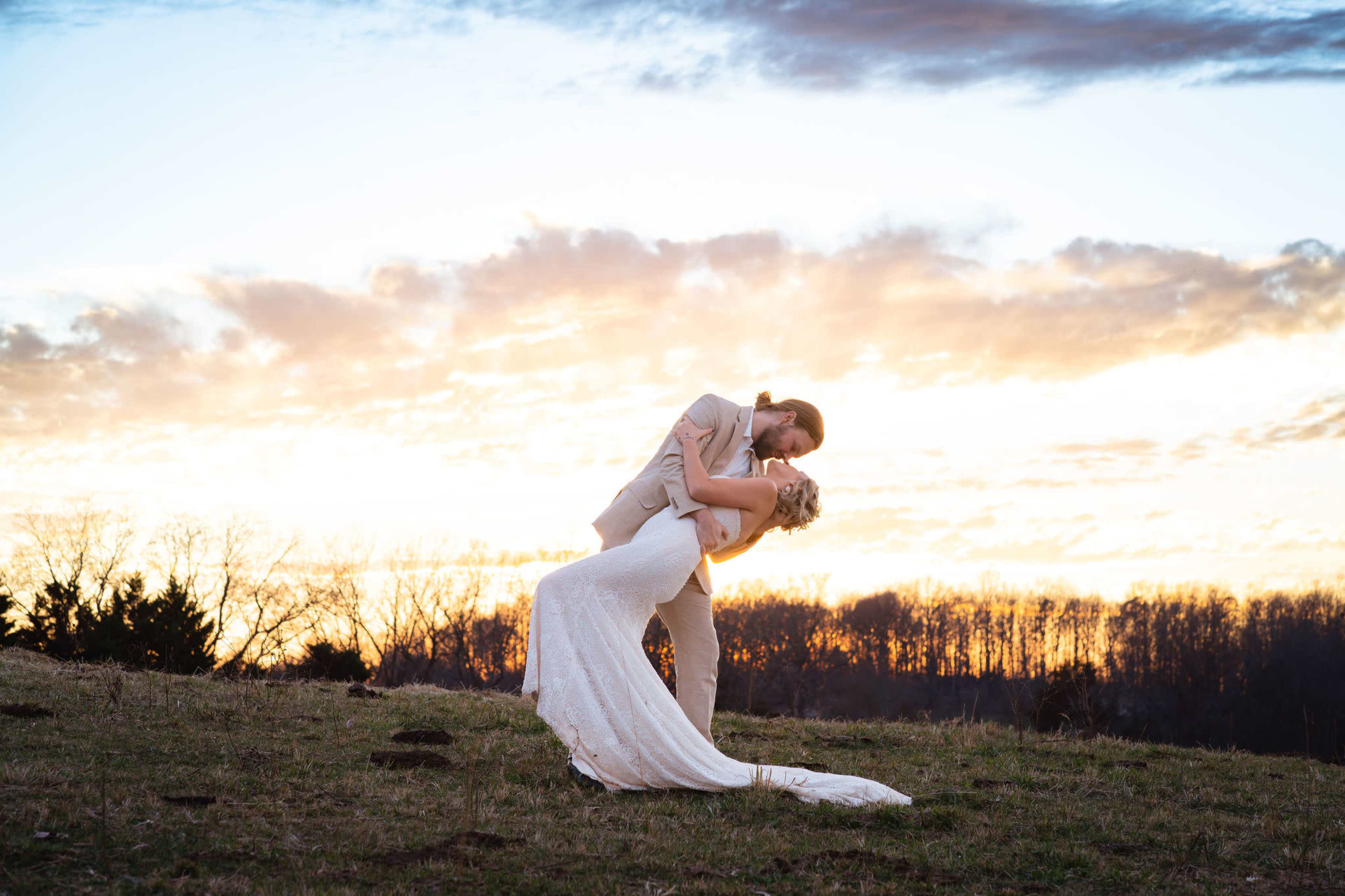 A couple in formal attire is captured mid-dip against a sunset backdrop with clouds.