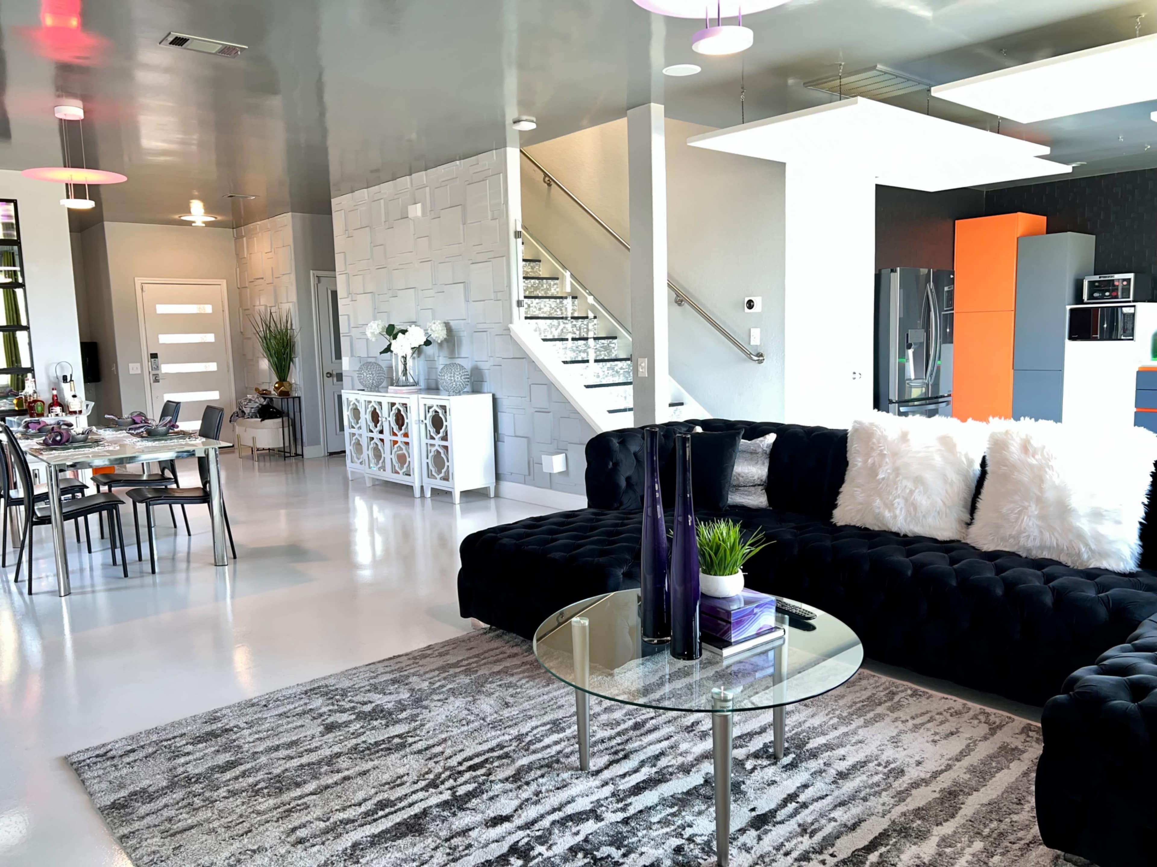 A modern living space featuring a black velvet sofa, a round glass coffee table, and an open dining area, with a staircase in the background.