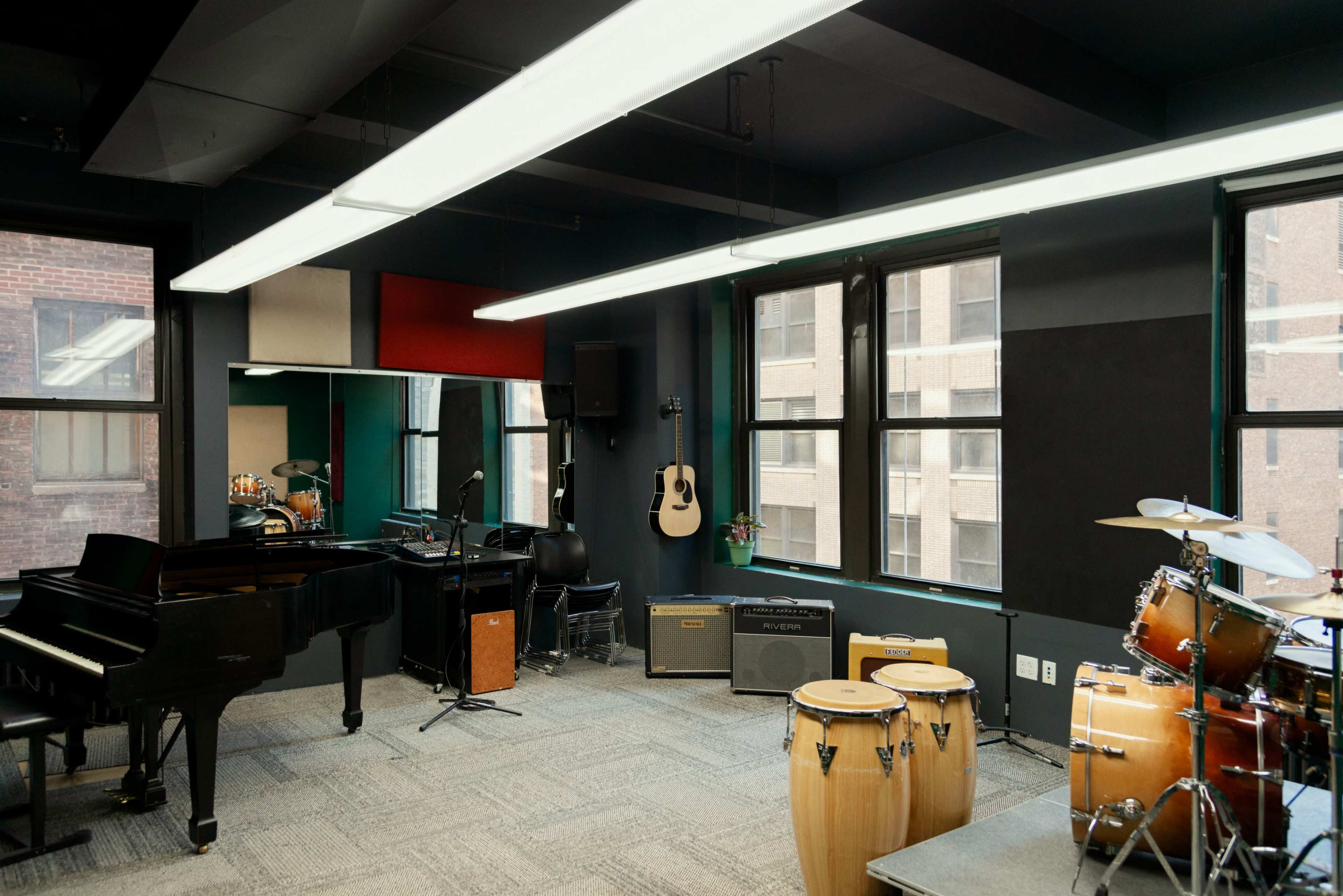 The room features a grand piano, drums, conga drums, an acoustic guitar on a wall, and large windows providing natural light.